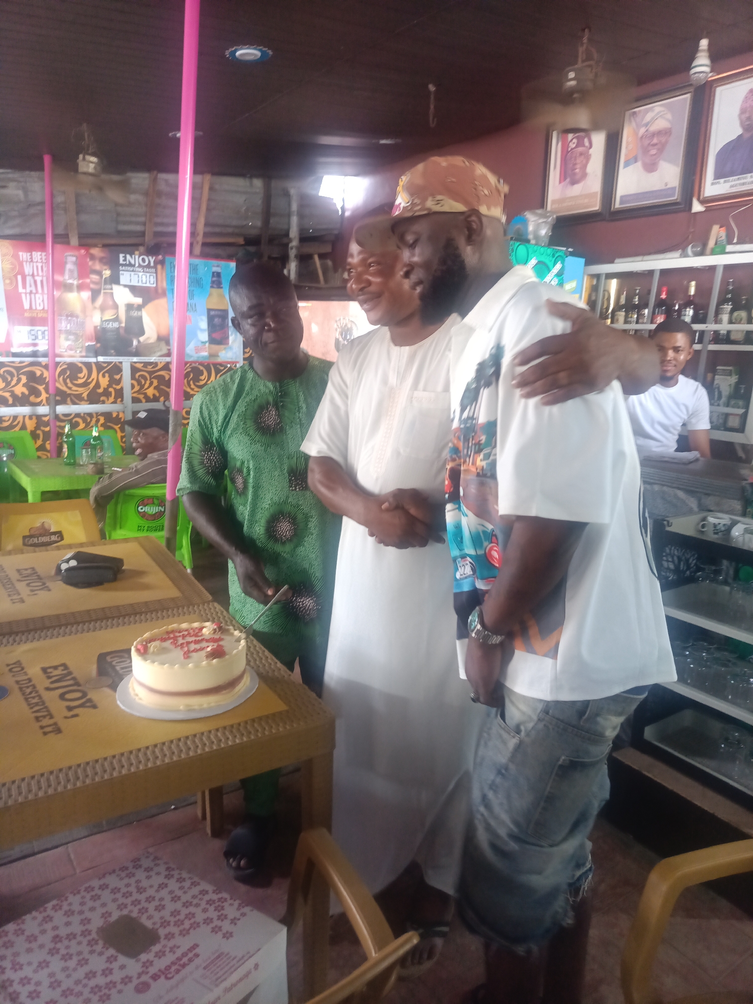 The image depicts an indoor scene, possibly a bar or restaurant, with several people present. In the foreground, a cake sits on a table, suggesting a celebration. Three men are prominently featured. One man, wearing a green patterned tunic, stands to the left. Another man, dressed in white, stands in the center, with his arm around the man to his right. This third man is wearing a white t-shirt with a colorful graphic and denim shorts, and he has a beard and is wearing a bandana. Behind them, a man in a white t-shirt is visible behind a counter, which is stocked with bottles. The background shows framed pictures on the wall and posters on a column. The overall impression is that of a casual gathering.