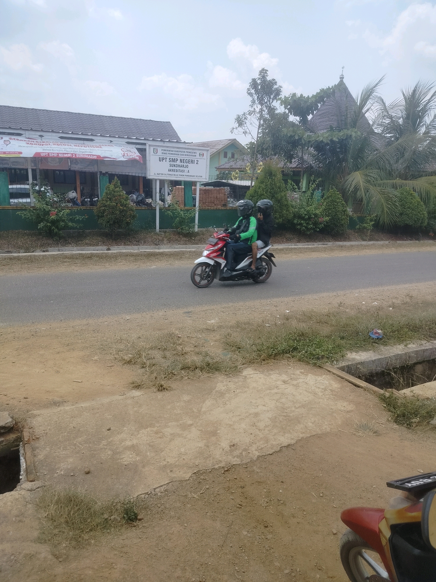 In Rejosari, Indonesia, a rural scene unfolds with a prominent school building on the left and a mosque-like structure in the background. A red and white motorcycle is captured in motion on the road, carrying two individuals wearing helmets and casual clothing. The rider, in a green shirt, is in front, while the passenger in a black shirt sits behind. The environment is characterized by a dusty roadside with sparse vegetation and a concrete drain. The sky is overcast, suggesting a potentially humid climate. The school building displays signage, including "UPT SMP NEGERI 2 SUKOHARJO," indicating it is a public junior high school. The mosque structure features a distinctive conical roof, common in traditional Indonesian architecture. The image appears to be taken during daytime.