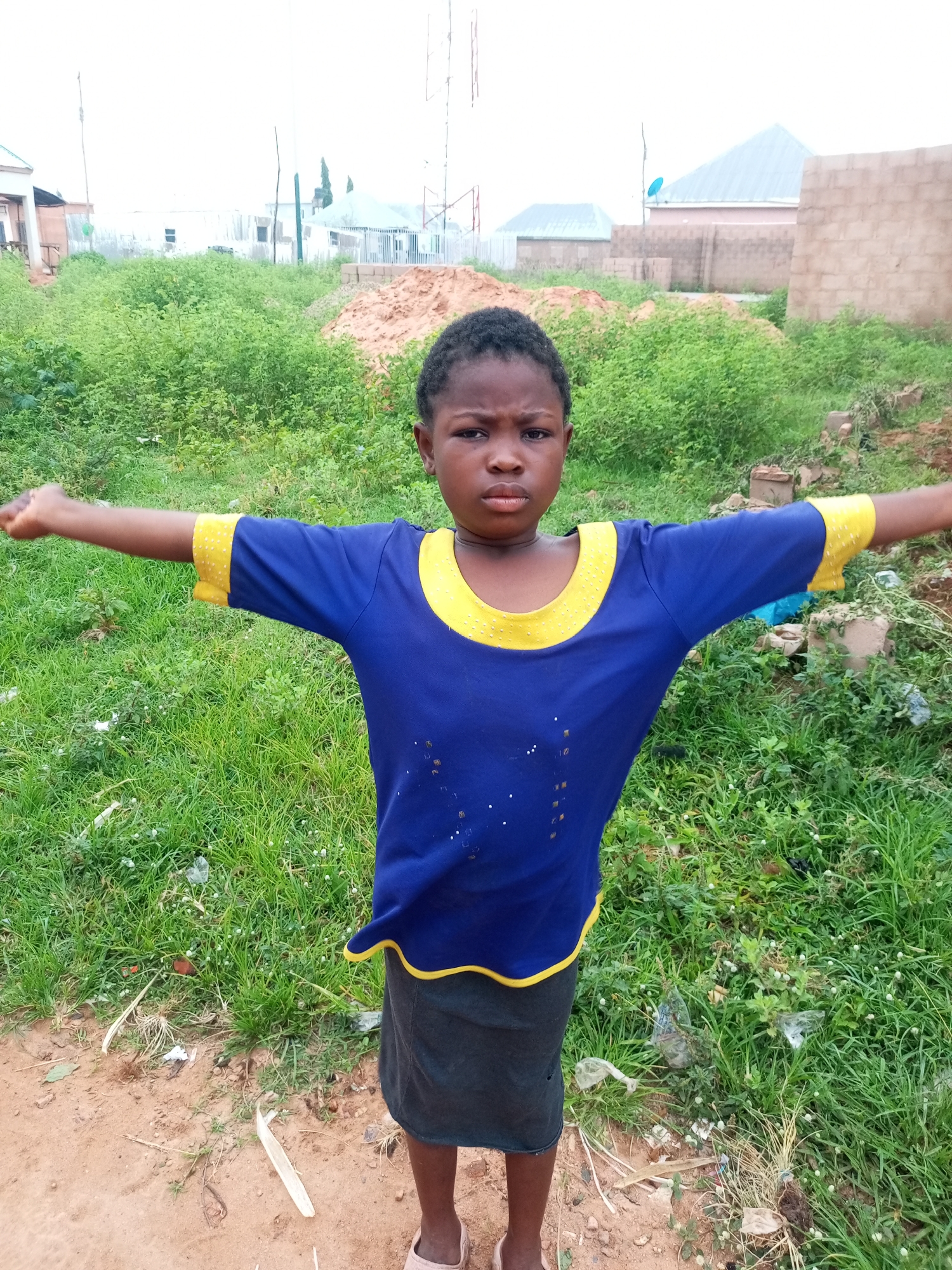 A young girl stands with her arms outstretched in a dirt and grass area, with buildings and construction materials in the background. The girl has dark skin and short, dark hair. She is wearing a blue t-shirt with yellow trim around the collar and sleeves, and a dark skirt. The ground is a mix of reddish-brown dirt and green grass, with some scattered debris, including plastic bags and dried leaves. In the background, there are unfinished buildings made of concrete blocks, a pile of sand or dirt, and some vegetation. The sky is overcast. The scene appears to be an outdoor area in a developing urban or semi-urban environment. The presence of construction materials suggests ongoing development or a construction site. The girl's pose, with arms outstretched, might indicate playfulness or a gesture of embrace. The overall setting suggests a humble living environment.