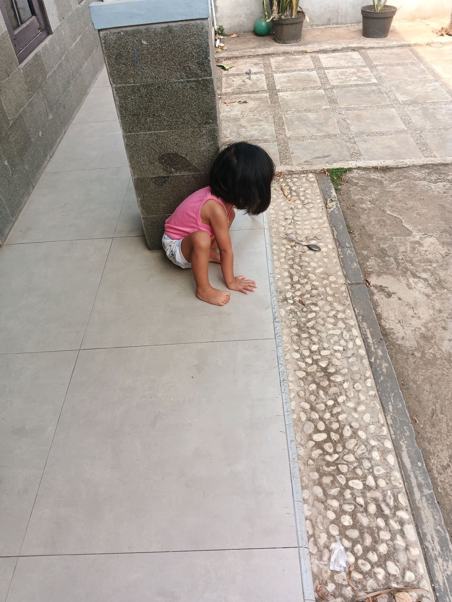 A young child, appearing to be a girl with dark hair, is sitting on a light gray tiled floor in a crouched position, leaning against a large, dark gray stone pillar. She is wearing a pink tank top and a diaper with a pattern. Her head is bowed, obscuring her face, and her hands are resting on the floor in front of her. The pillar is positioned at the corner of a building with a rough, gray stone facade. To the right of the pillar, a textured pathway is visible, made of concrete with embedded small white stones. Beyond this pathway is a rougher concrete ground with some dry leaves and debris. In the background, two potted plants are visible on the concrete ground. The overall scene suggests an outdoor area, possibly a courtyard or patio, with elements of urban infrastructure. The location is specified as Kadu, Indonesia.
