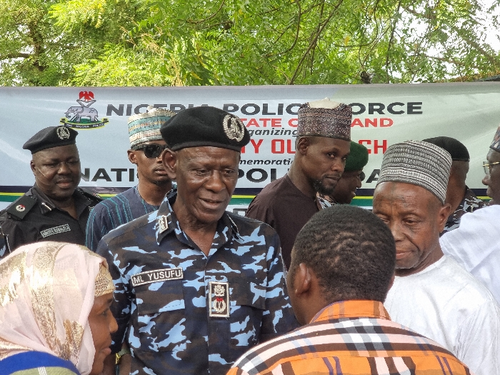 The image shows a group of people standing in front of a banner that reads "NIGERIA POLICE FORCE STATE COMMAND ORGANIZING MY OWN CH MEMORATIO POLI." The banner is located in Maiduguri, Borno State, Nigeria. The group is composed of mostly men, some are wearing traditional Nigerian clothing and headwear, including a few in police uniform. In the center of the group is a man in a police uniform and cap, speaking to the people. The man is identified by a nametag as "ML YUSUFU". The individuals are interacting in a gathering setting, possibly a community event or gathering. There are trees and greenery in the background.