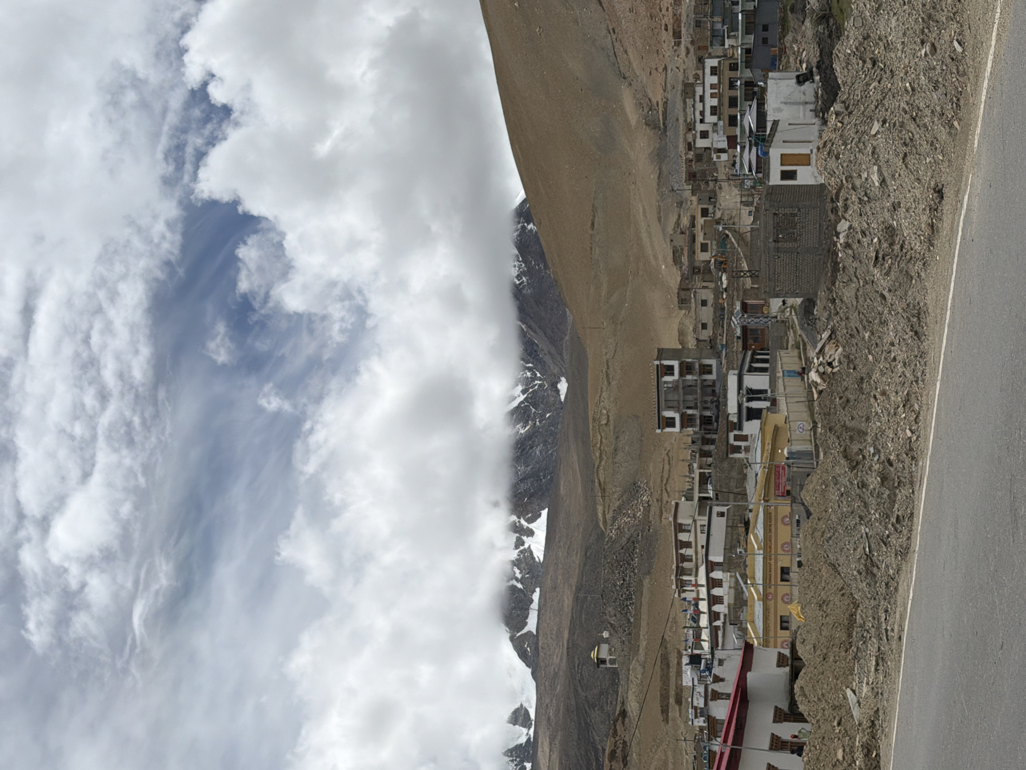 This is a medium shot photograph taken from a low angle, looking up at the village of Karzok, India, situated in the Ladakh region. The scene is dominated by the stark, brown, mountainous landscape and a cloudy sky. In the foreground, a paved road runs horizontally across the bottom of the frame, with a pile of dirt and debris along its edge.
The middle ground features a cluster of buildings that constitute the village. These structures are varied in design and construction, with some appearing to be traditional Tibetan architecture featuring white walls and darker, flat roofs, while others are more modern, with concrete and brick elements. Several buildings have multiple stories. Flags can be seen flying from some of the buildings. Among the buildings, there is a yellow structure with text on it, which appears to be a community center or hall.
In the background, a massive mountain range rises, its peaks covered in snow. Clouds shroud the upper parts of the mountains, obscuring some of the detail. To the left, on a hillside overlooking the village, a small, white structure with a golden dome, possibly a stupa or a small temple, is visible.
The overall atmosphere is one of rugged, high-altitude living. The lighting is diffused due to the cloudy conditions, resulting in soft shadows and a somewhat muted color palette. No people are clearly discernible in the image, though there might be figures in the shadows of the buildings, their presence is not distinct. The environment suggests a remote and arid location.
