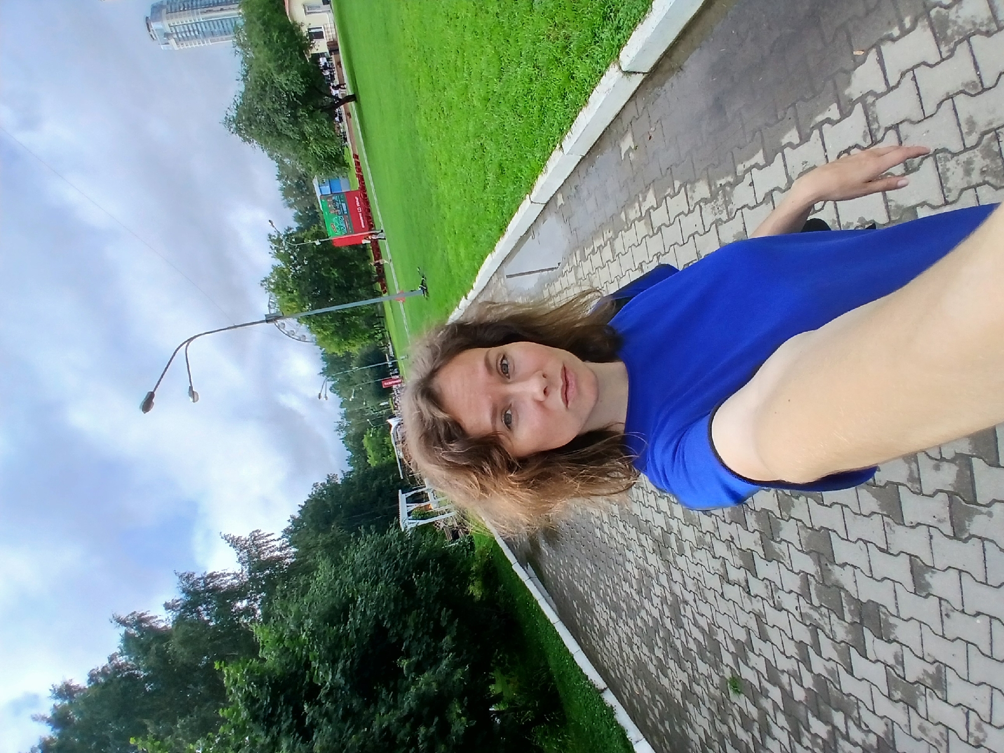 A woman with curly brown hair, wearing a blue t-shirt, is taking a selfie on a paved walkway in a park. The walkway is wet, suggesting it may have rained. To the left of the walkway is a grassy area bordered by trees. In the background, there is a Ferris wheel, a billboard, a tall building, and other park-goers. The sky is cloudy. The location is likely a park in Yekaterinburg, Russia, possibly the Mayakovsky Park or the Park of the XXII Parts Congress, given the presence of a Ferris wheel and a tall modern building visible in the distance.