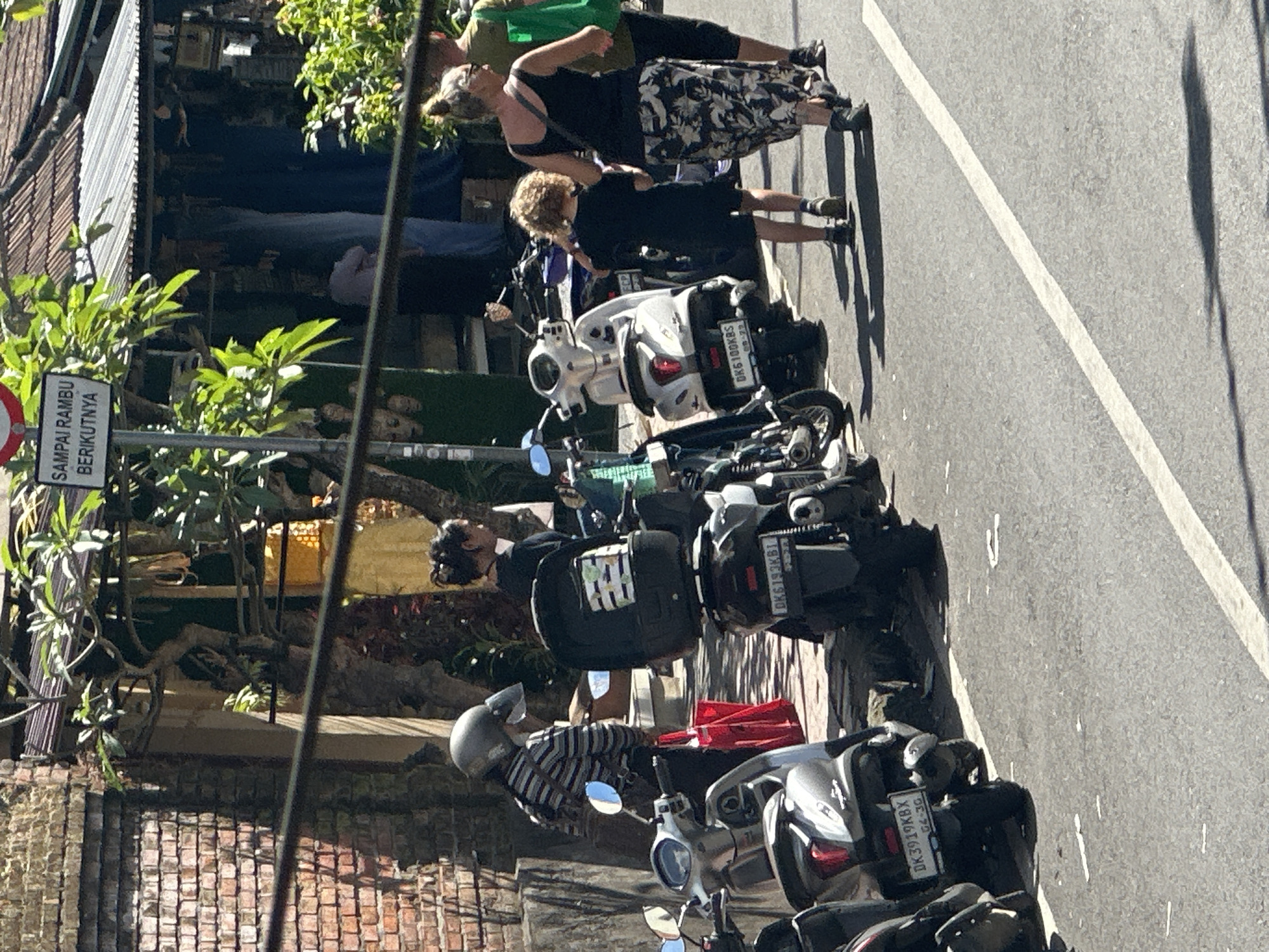 In Ubud District, Indonesia, on a sun-drenched street, a variety of parked scooters are lined up along a paved roadway. To the left, a brick wall forms part of a building, while to the right, a shop with a corrugated metal roof displays various items, including textiles and masks. The scene is bustling with activity. A woman wearing a black tank top and a patterned skirt walks away from the camera, her hair in a bun. Beside her, a young child with curly hair, dressed in a black t-shirt and shorts, walks in the same direction. Another woman, in the foreground, wearing a black top and a black and white floral skirt, walks further away. Further back, a person in a striped long-sleeved shirt and helmet, holding a red bag, stands near a silver scooter. A woman with her hair in a bun, wearing a black top, is partially visible behind the person in the helmet. Several scooters are parked along the street, some with storage boxes on the back, and their license plates are visible, such as "DK6100KBS" and "DK3919KBX." Trees with lush green leaves overhang the scene, providing shade. A sign with text in Indonesian, "SAMPAI RAMBU BERIKUTNYA," is mounted on a pole. The overall impression is one of a vibrant street scene in a tropical locale.