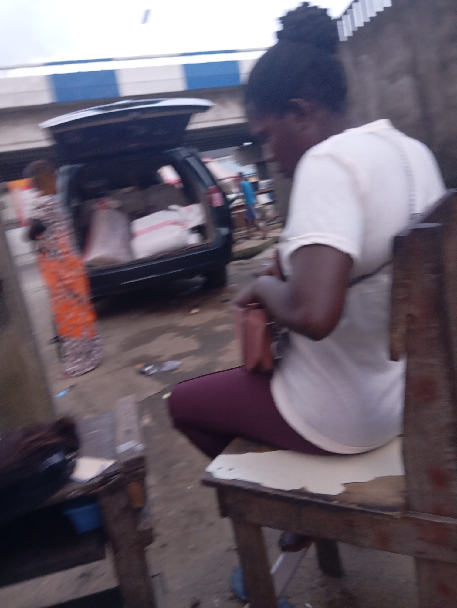 In Port Harcourt, Nigeria, a blurred image captures a street scene. In the foreground, a woman in a white t-shirt and burgundy leggings sits on a wooden chair. She holds a pink purse in her lap. To her left, another woman in an orange and grey patterned dress stands near the open trunk of a dark-colored SUV, which is loaded with sacks. In the background, a person in a blue shirt walks away from the vehicle. The environment appears to be an outdoor market or roadside area, with concrete ground and hints of buildings and a striped overpass.