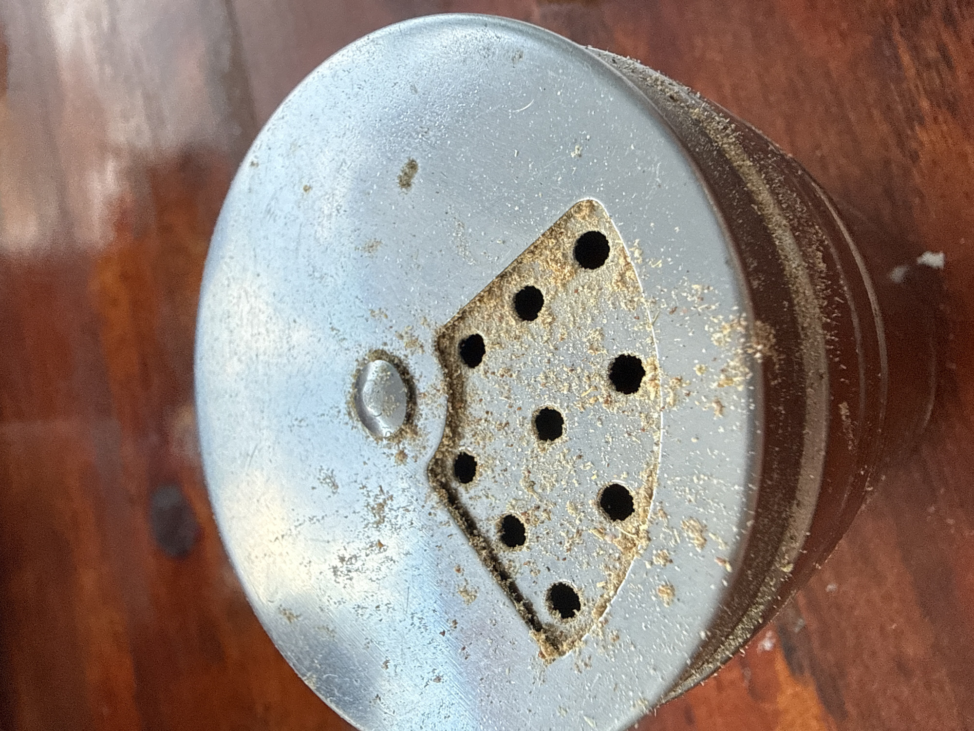 The image shows a close-up, overhead view of the metal cap of a spice shaker, resting on a wooden surface. The cap is circular with a textured, slightly worn metallic finish. A triangular section of the cap has been cut out to create a series of small holes, some of which are filled with a light-colored granular substance, likely dried spices. This triangular area is also visibly dusty. The rest of the cap has a raised, circular indentation in the center, possibly for a different shaker setting.
The wooden surface beneath appears to be a table or countertop, with a warm, reddish-brown hue and visible wood grain. The lighting suggests an indoor setting, possibly during the daytime, as there are no strong shadows indicating artificial light. There is no text visible in the image. The overall impression is of a common household item, perhaps in use or storage, showing signs of age and use. The location context of Sozopol, Bulgaria, does not provide any specific visual cues within the image itself.
