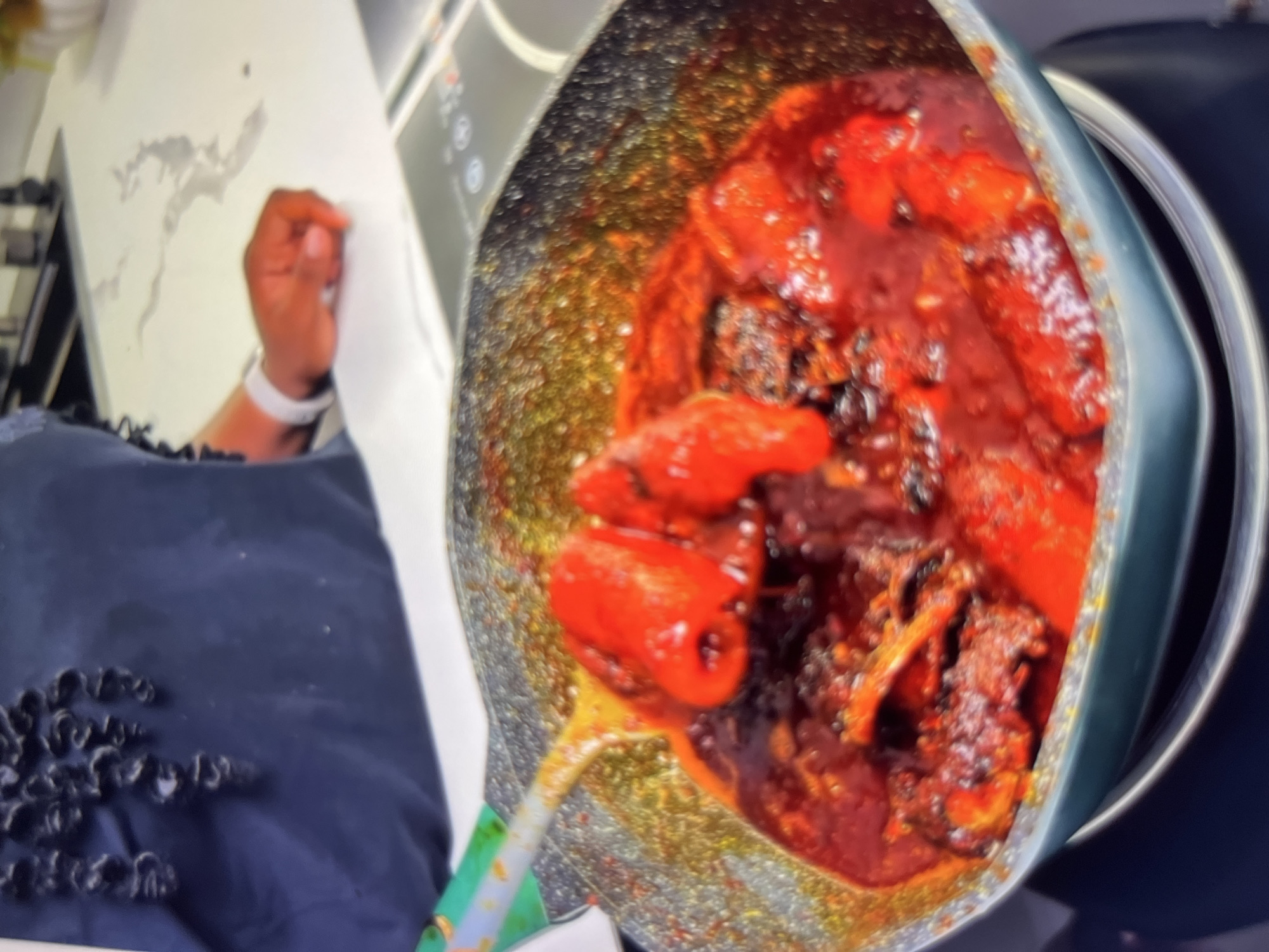 An overhead, close-up shot captures a pan of food bubbling with a rich, red sauce, containing what appear to be chunks of meat and possibly tomatoes. A green-handled spoon is partially submerged in the sauce, suggesting active cooking. The pan is dark gray with a speckled texture. In the background, a person is partially visible, wearing a dark blue garment with a textured pattern. Their arm rests on a white countertop, and a white smartwatch is visible on their wrist. The setting appears to be a kitchen, with a stovetop and countertop elements visible. There are no specific landmarks or indications that this scene is located in Abuja, Nigeria.