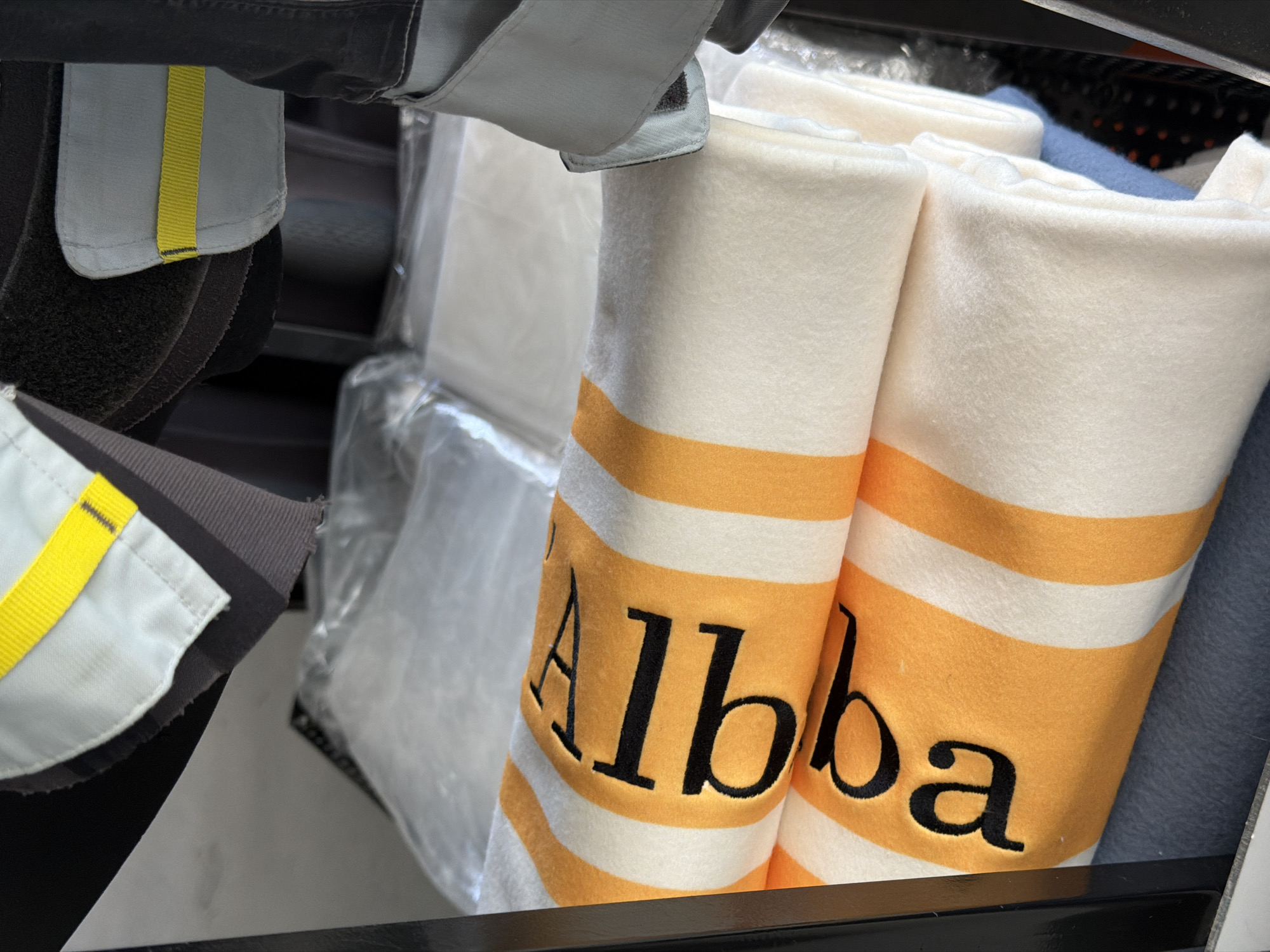 The image displays an organized collection of textile items, likely within a storage unit or a utility cart in an interior setting. In the upper portion, fragments of what appear to be functional gear are visible, including a light grey fabric section with a prominent yellow webbing stripe, and a darker, textured material, possibly black or dark grey, suggestive of a padded or Velcro component. The central focus is on two identically patterned blankets, folded and stacked. These blankets feature a repeating design of alternating white and orange horizontal stripes. Embroidered or printed in black serif font on the orange stripes of the top blanket is the word "Alba," readable if oriented correctly or interpreted as reversed text ("qIbA" is visible). Below these, a solid blue fleece-like fabric is partially visible, and further down, a dark textile with a pattern of light-colored dots or specks can be seen. In the background, on what appears to be an upper shelf or compartment, several rectangular white items are individually sealed in clear plastic bags, indicating a need for sterility or protection. The overall scene suggests a functional environment, such as a supply room, a medical facility, or an emergency service vehicle. The image contains no visual information, such as landmarks, signage, or specific cultural identifiers, to indicate its location in the city of Saint Petersburg, Russia, or any other particular geographical area.