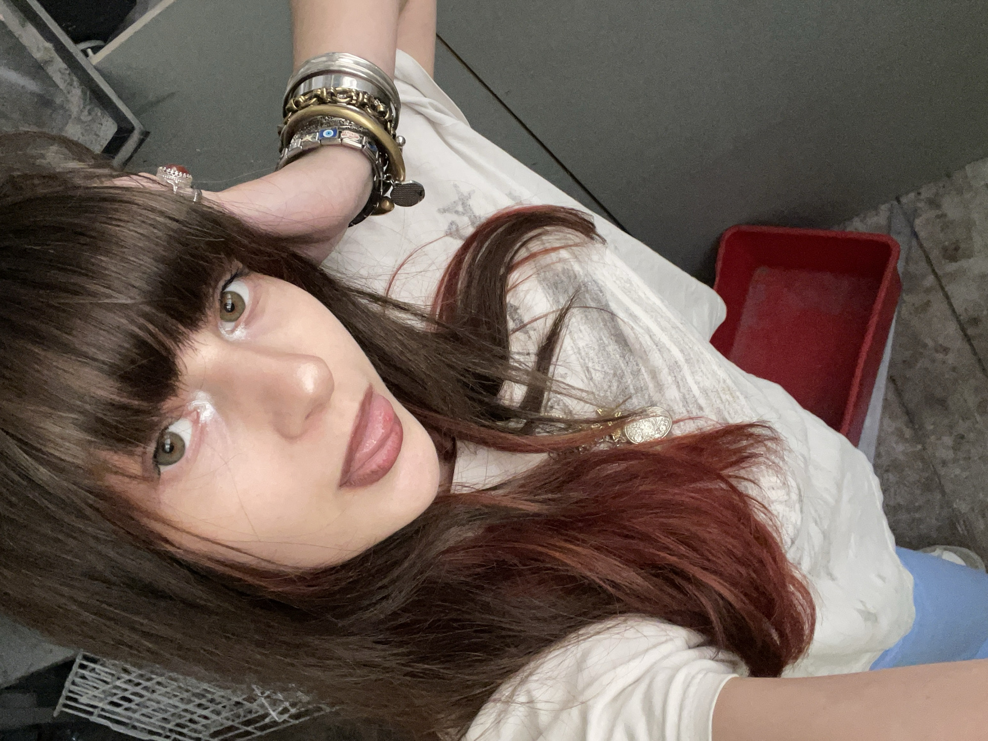 A selfie taken from a high angle shows a person with brown hair, some of which is dyed red, brown eyes, and fair skin. They are wearing a white top and several bracelets on their left wrist, including one with an evil eye bead. A ring is visible on a finger of the left hand. The background includes a gray wall and a red plastic bin. The location is unknown and cannot be determined from the image.