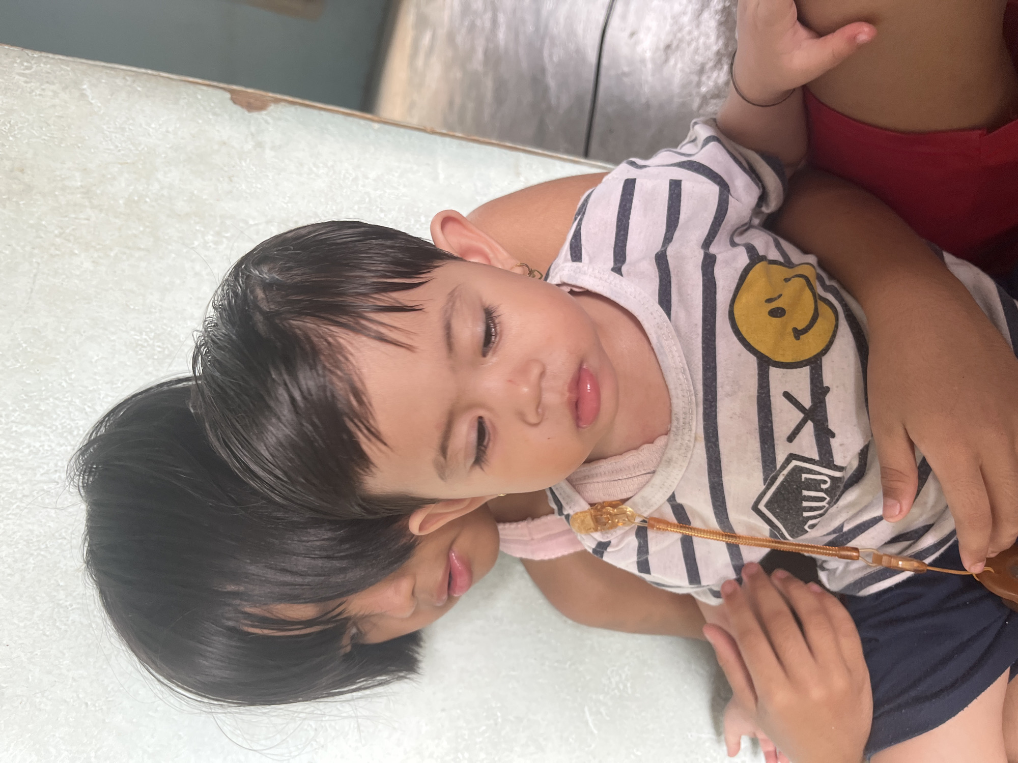Two young children, likely siblings, are seated closely together against a textured, pale wall. The child in the foreground, appearing to be a toddler, is held by the child behind. The toddler, facing forward with their head slightly tilted down, wears a striped shirt with a smiley face graphic. The older child, whose face is partially obscured by their dark hair, is wearing a pink top and is positioned behind and to the left of the toddler. Their hands are visible, one gently resting on the toddler's chest and the other on their thigh. The scene appears to be indoors, suggested by the wall texture and what appears to be a concrete floor in the background. The lighting is soft and natural, indicating daytime. This image was taken in a residence in Meliau Hulu, Indonesia.
