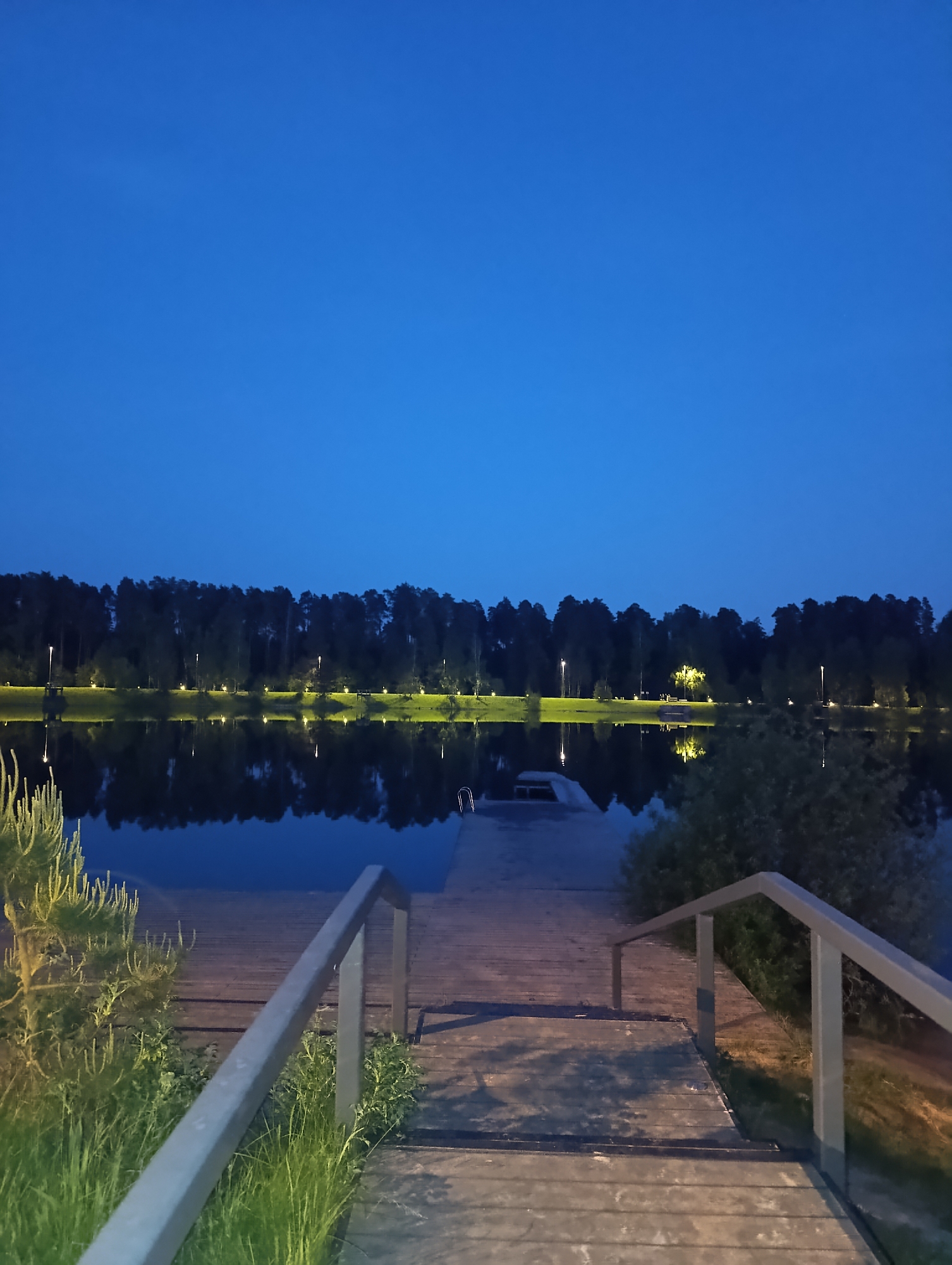 A tranquil outdoor scene captures a wooden staircase leading down to a tranquil lake, likely at dusk or dawn. The sky overhead is a deep, clear blue, transitioning into a darker horizon where a thick line of trees stands silhouetted against the fading light. The trees reflect softly in the still water of the lake, creating a mirror-like effect, only broken by the slight ripples on the water's surface. Along the tree line, lights are placed, evenly spaced. The staircase is made of planks of wood, with metal railings on either side. The wood shows signs of weathering, but appears well-maintained. Near the base of the stairs, patches of greenery fill the edges. The overall scene is peaceful, suggesting a park or recreational area within the city of Unknown, Unknown.