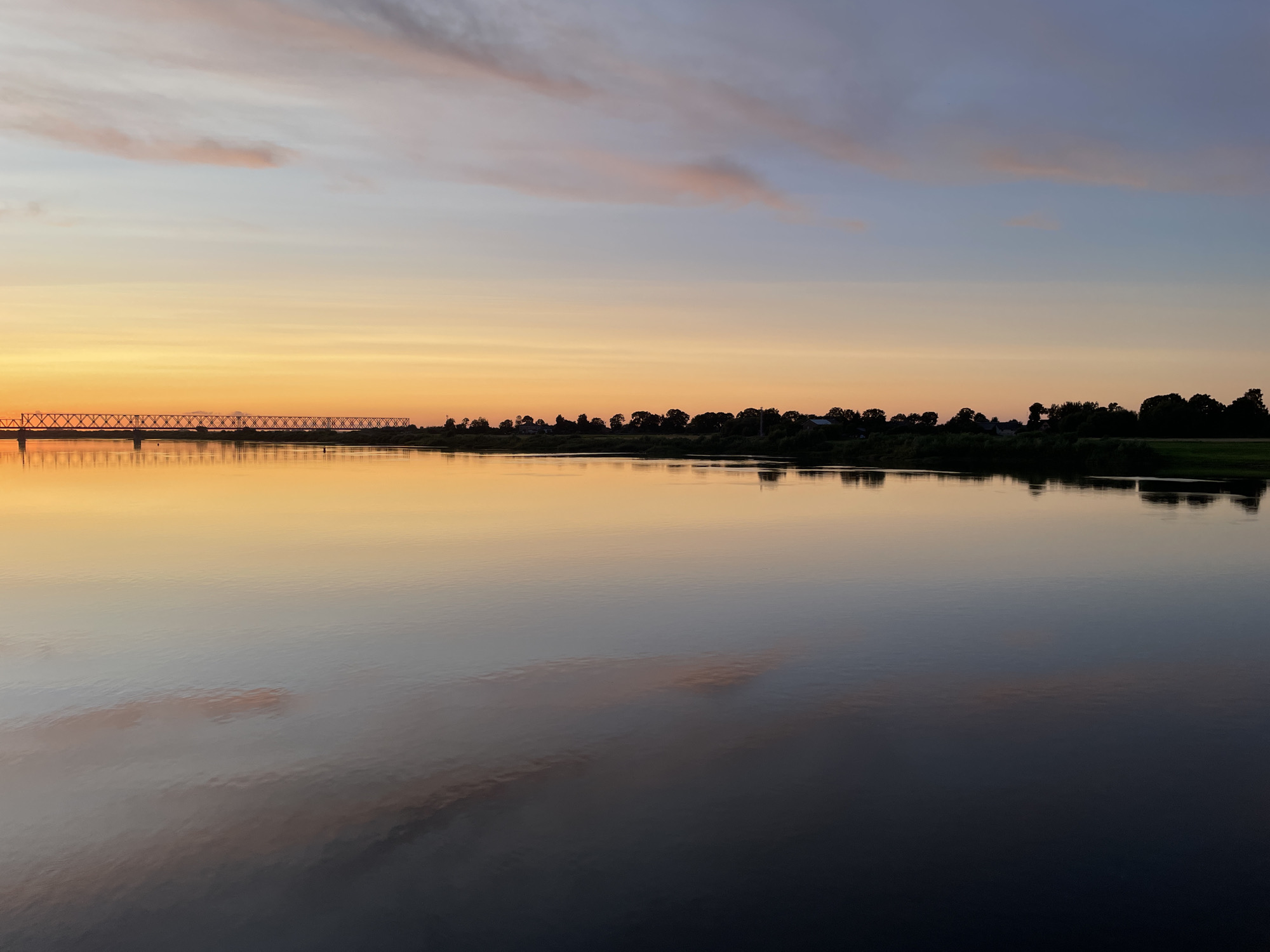 A serene, wide river reflects the soft hues of a twilight sky over Sovetsk, Russia. A long, metal truss bridge spans the river in the distance, its structure silhouetted against the fading orange and pink glow. The opposite bank is lined with a dense band of dark trees and scattered buildings, their shapes softened by the low light. The water's surface is remarkably still, mirroring the sky and the distant landscape with perfect clarity, creating a sense of peaceful symmetry. The sky transitions from a warm orange near the horizon to cooler blues and purples overhead, with wispy clouds catching the last light of the day. The overall impression is one of tranquility and natural beauty at the close of day in this Russian city.