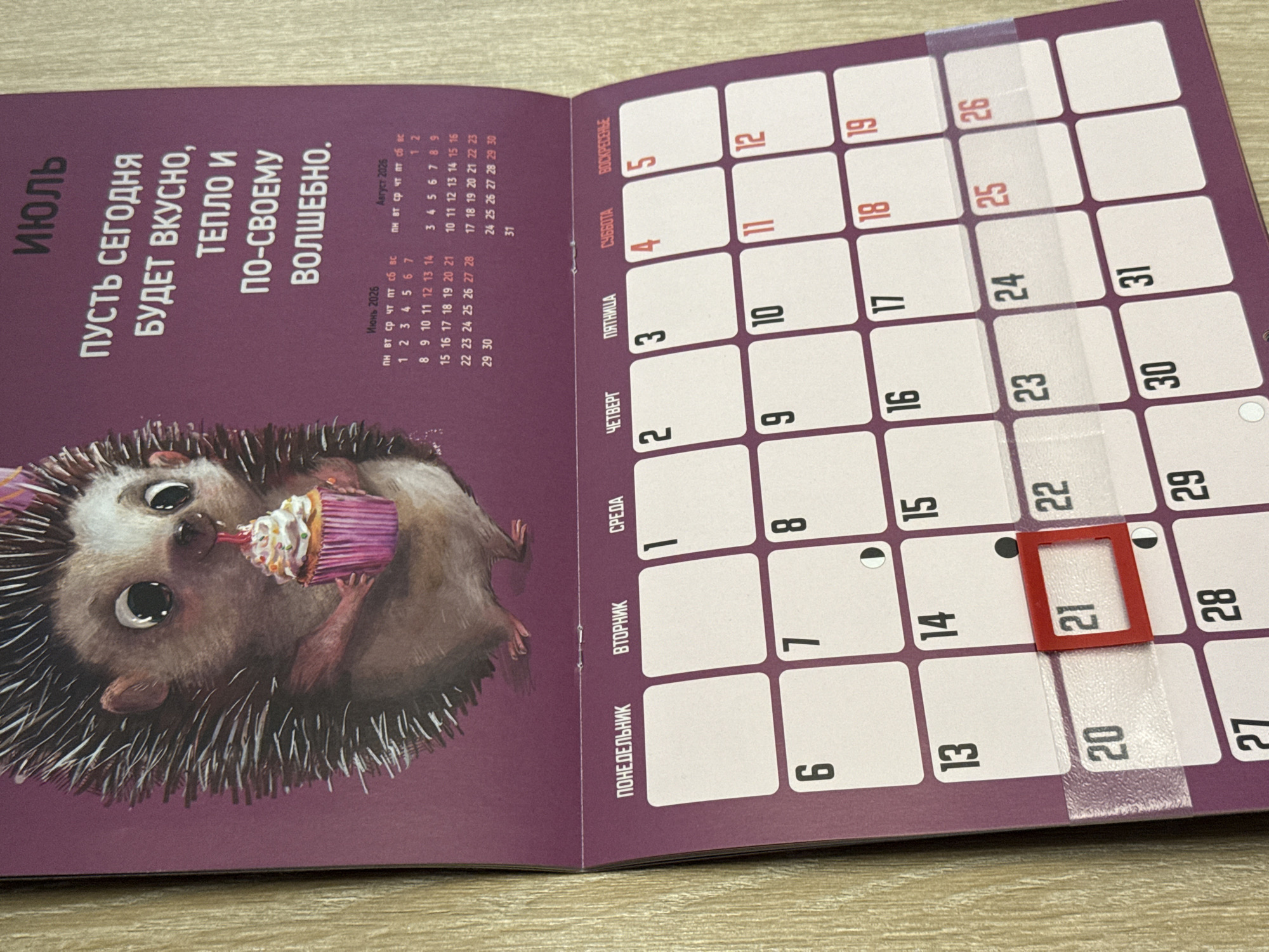 The image shows a close-up of a calendar page for July. The calendar features a whimsical illustration of a hedgehog holding a cupcake. The calendar grid is visible below the illustration, with days of the week labeled in Russian: "ПОНЕДЕЛЬНИК" (Monday), "ВТОРНИК" (Tuesday), "СРЕДА" (Wednesday), "ЧЕТВЕРГ" (Thursday), "ПЯТНИЦА" (Friday), "СУББОТА" (Saturday), and "ВОСКРЕСЕНЬЕ" (Sunday). The current date, the 21st of July, is highlighted with a red frame. The calendar also includes small icons indicating lunar phases on some days. The surrounding environment appears to be a wall with a wooden texture visible on the right side. There is no information in the image to suggest a location in Lyubertsy, Russia.