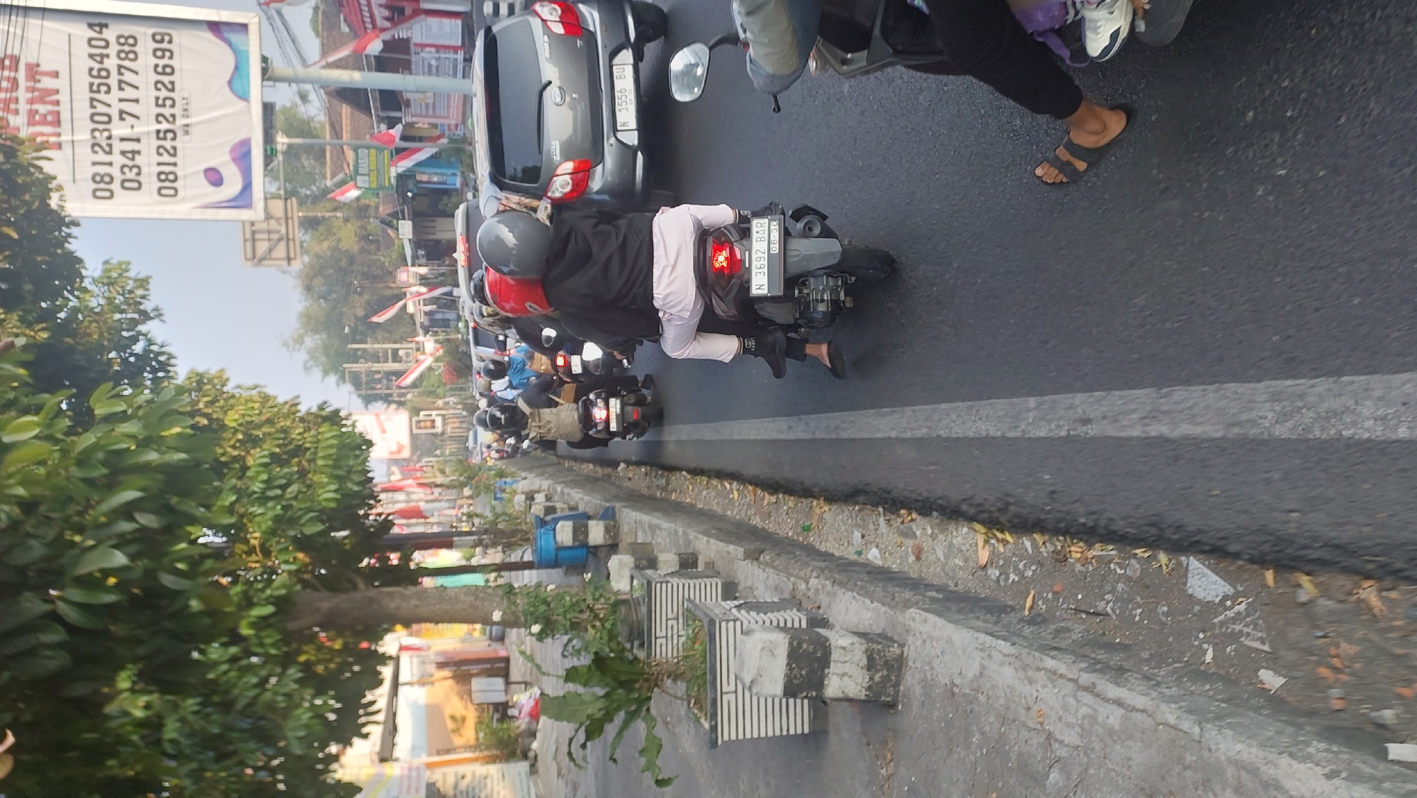 The image depicts a street scene in Malang, Indonesia, likely during daylight given the bright sky and shadows. The perspective is from the rear of a motorcycle, looking forward. In the foreground, a person is riding a motorcycle, wearing black pants and sandals, and a black hoodie. The motorcycle has a license plate that reads "N 3692 BAR" and displays the date "06.30". Another motorcycle is visible to the left, with a rider wearing a red helmet.
Further down the road, traffic is moderate, with several other motorcycles and cars visible. Some of the motorcycles have riders wearing helmets. A grey car with a license plate "N 3556 BU" is in the right lane. The environment includes a concrete sidewalk with planters containing green foliage, and a drainage ditch. Buildings with Indonesian flags are visible in the background, suggesting a national holiday or celebration. A billboard with text and a phone number is also present on the right side of the road. The road surface is asphalt, and there are painted white lines indicating lanes. The overall impression is of a typical urban street with traffic in Indonesia.