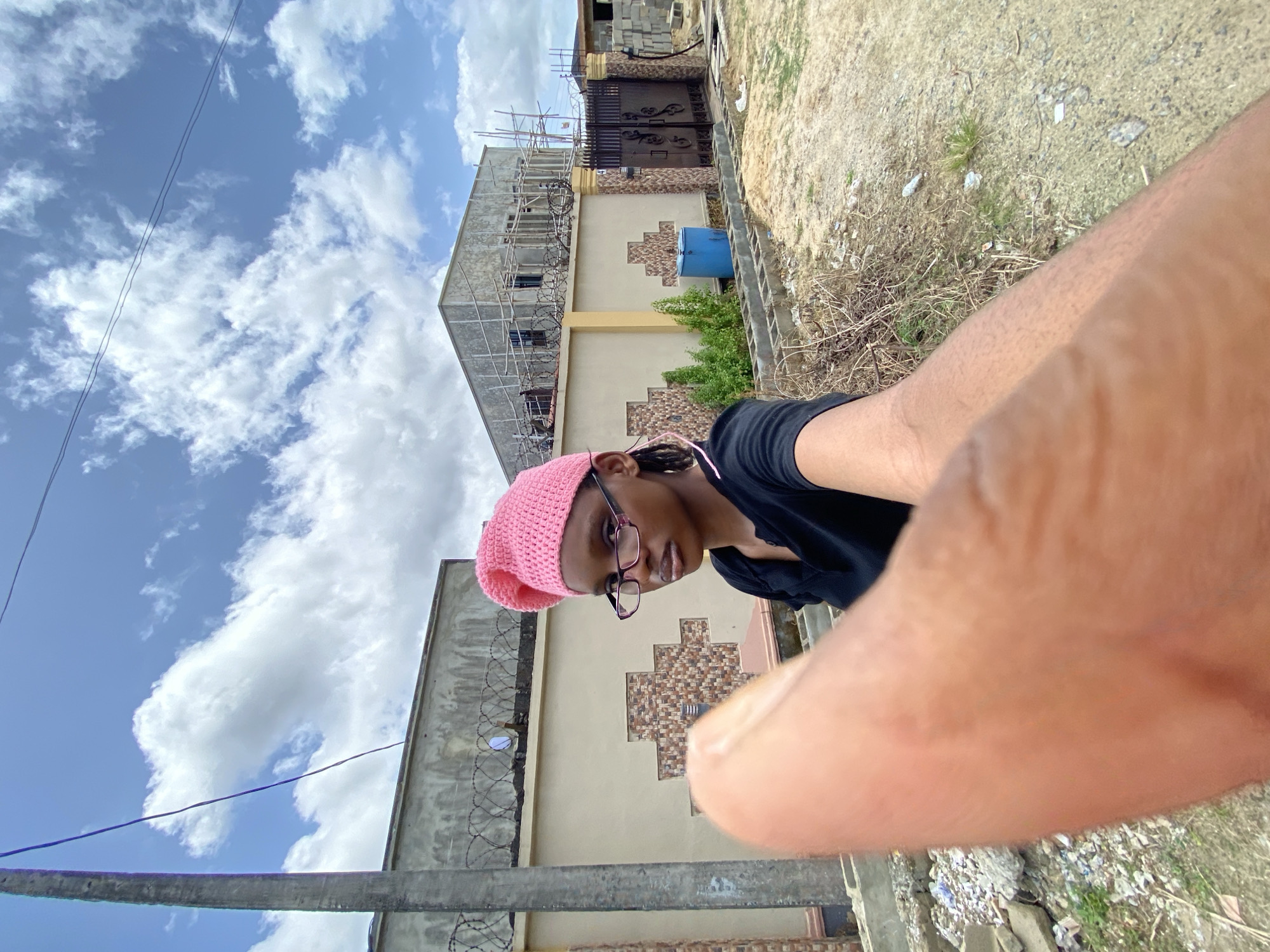 A selfie-style, rotated, wide-angle shot captures a person with brown skin wearing a pink knit hat and glasses. They are dressed in a black shirt and look downwards at the camera. The foreground is obscured by a large, blurred forearm and hand.
Behind the person, a partially constructed building is visible, flanked by a light-yellow building with tan and brown patterned accents. Both buildings are topped with barbed wire. A blue water tank is situated near the building under construction. The ground is covered in dry vegetation and debris. The sky is visible in the background, featuring white clouds against a blue sky.
The location appears to be an urban or suburban area with ongoing construction. The exact city and region is unknown.