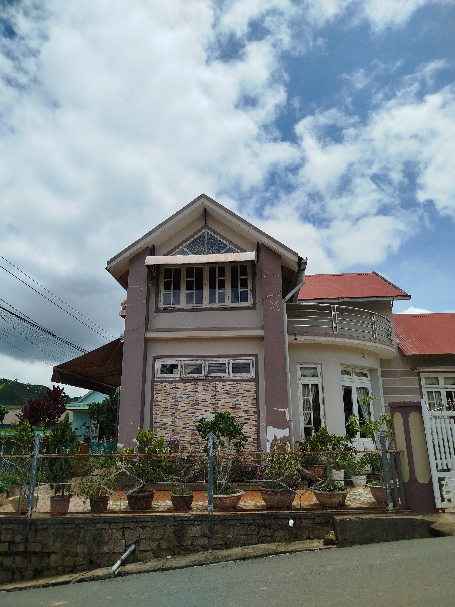 This is a full shot, eye-level, exterior photograph of a two-story house with a red-tiled roof, located in the city of Xuân Sơn, Vietnam. The house has a light brown and white facade with brown trim around the windows and doors. The front of the house features several windows and a balcony on the second floor, with a curved metal railing. The ground floor has a large, arched doorway with white double doors and is lined with potted plants. A white metal gate is visible on the right side of the house. The house is surrounded by a fence with a stone wall at its base, and more potted plants are arranged along the fence. The sky is blue with scattered white clouds, and a green hill is visible in the background. The street in front of the house is paved with asphalt.