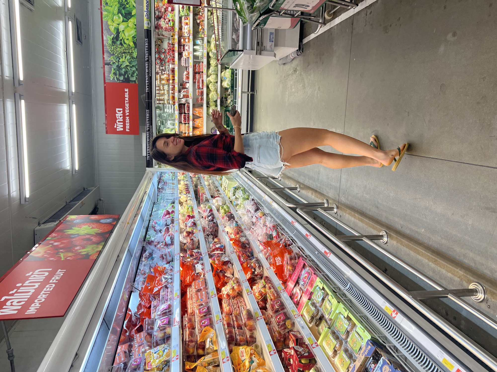A young woman with long brown hair stands in an aisle of a supermarket. She is wearing a red and black plaid shirt, denim shorts, and yellow flip-flops. She is holding a small object in her right hand.
Behind the woman are refrigerated displays filled with produce, including fruit and vegetables. Signage in Thai and English is visible, indicating "Imported Fruit" and "Fresh Vegetable." The supermarket aisle is brightly lit with fluorescent lighting. The location is likely in Baan Bo Phut, Thailand, given the Thai signage.
