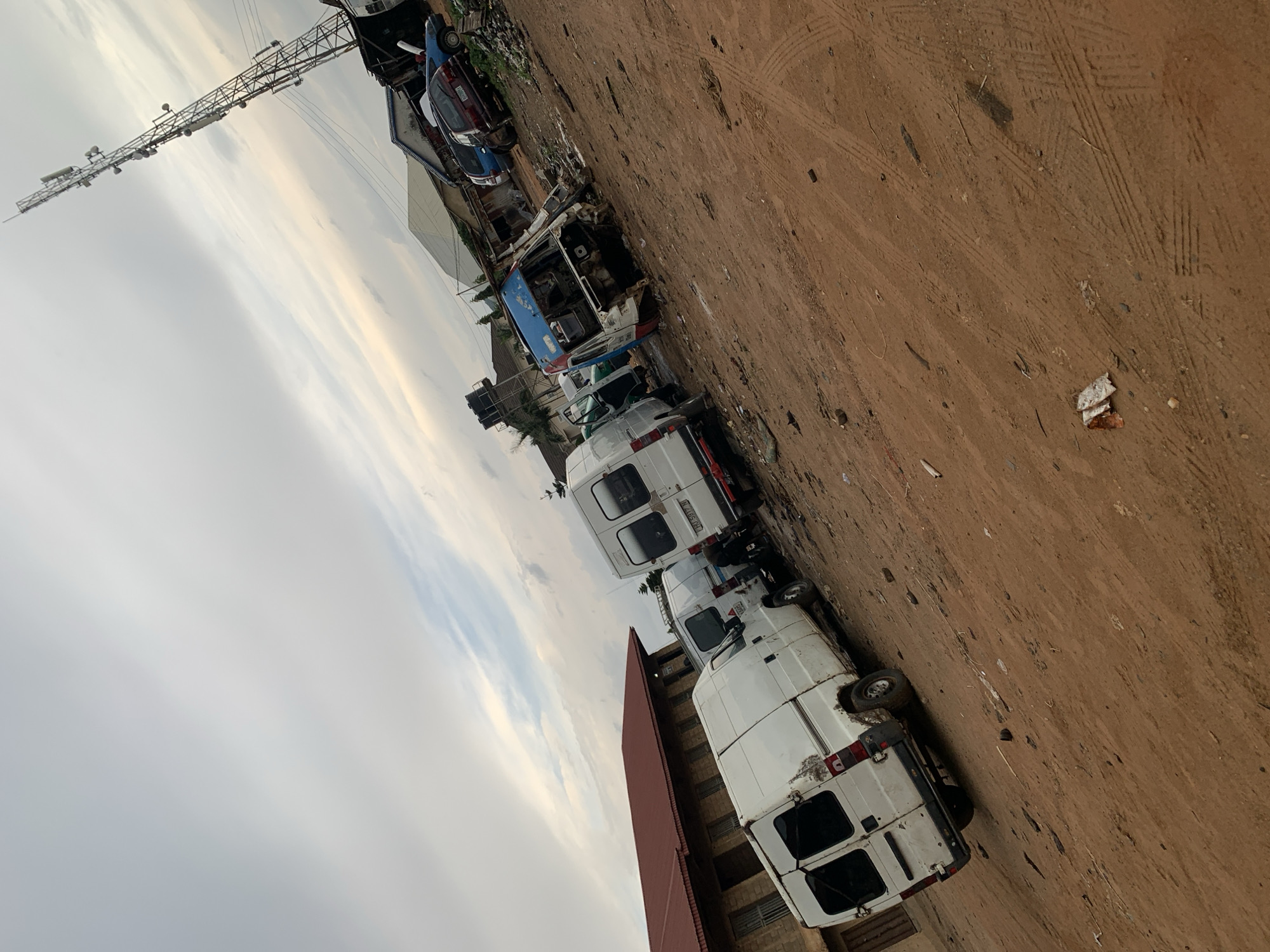 In Rafin Par, Nigeria, an outdoor scene depicts a collection of vehicles, predominantly white vans, parked on a dirt lot under an overcast sky. The vehicles are a mix of operational and seemingly abandoned. In the foreground, several white panel vans are visible, one of which is angled towards the viewer and shows signs of wear. To the right of these vans, a heavily damaged blue and white van is prominent, its front end dismembered. Further back, other vehicles including a dark-colored sedan and a blue car are parked. A large telecommunications tower dominates the background, alongside various buildings and structures, suggesting an urban or semi-urban environment. The ground is covered in reddish-brown dirt with tire tracks visible. Scattered debris is present throughout the lot.