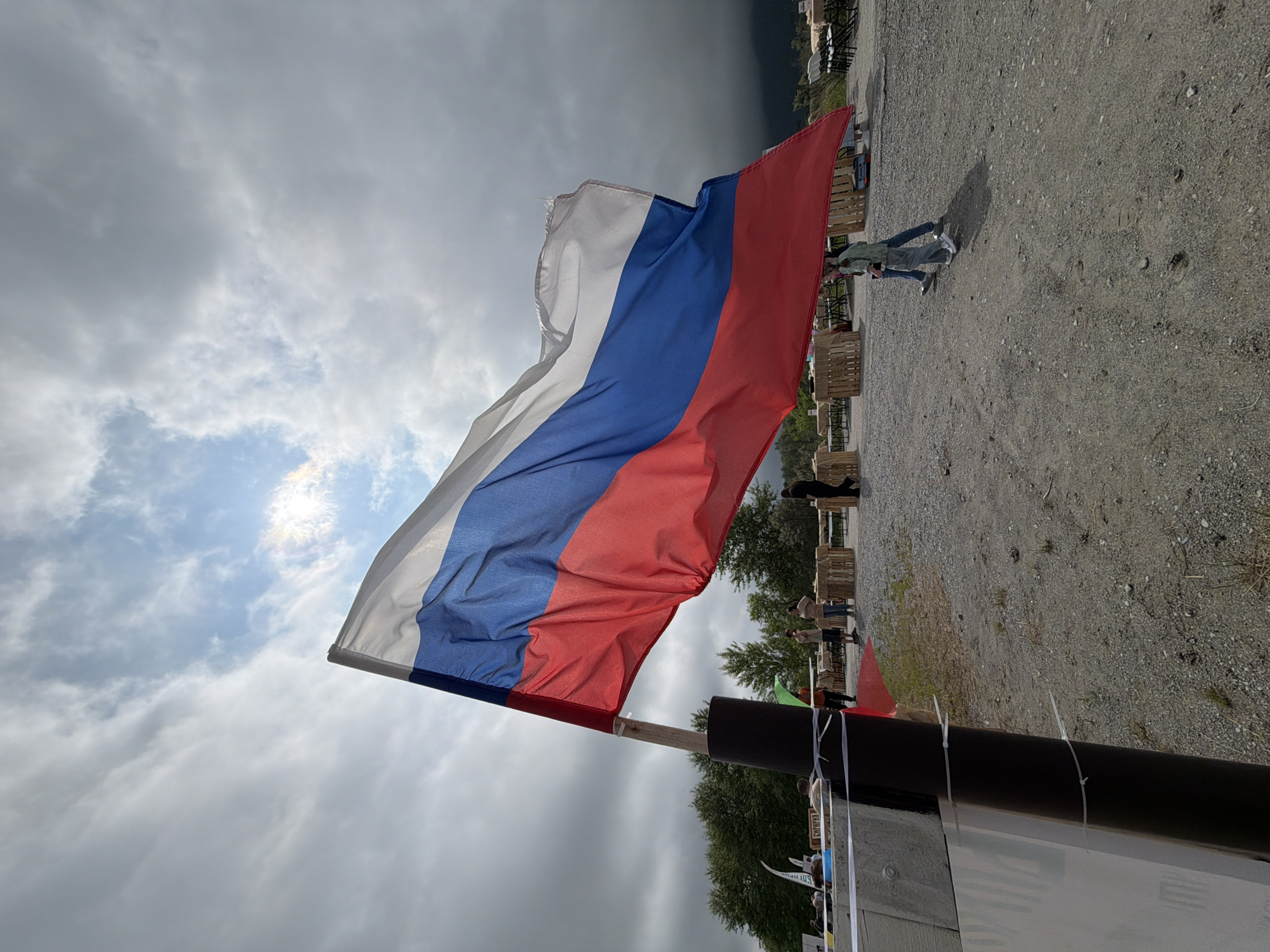 An outdoor scene features a large Russian flag prominently displayed, waving in the wind against a cloudy sky with patches of sunlight. The flag, composed of white, blue, and red horizontal stripes, is attached to a wooden pole wrapped in dark material. In the background, several people are visible, some standing near wooden crates and tables, suggesting an event or gathering. Trees and hills form the distant landscape. The ground is covered with gravel and dirt, and some white tape barriers are present in the foreground. The scene appears to be in an open, possibly rural or park-like setting, with a few structures and seating areas visible. The overall impression is one of a public outdoor event under a dynamic sky.