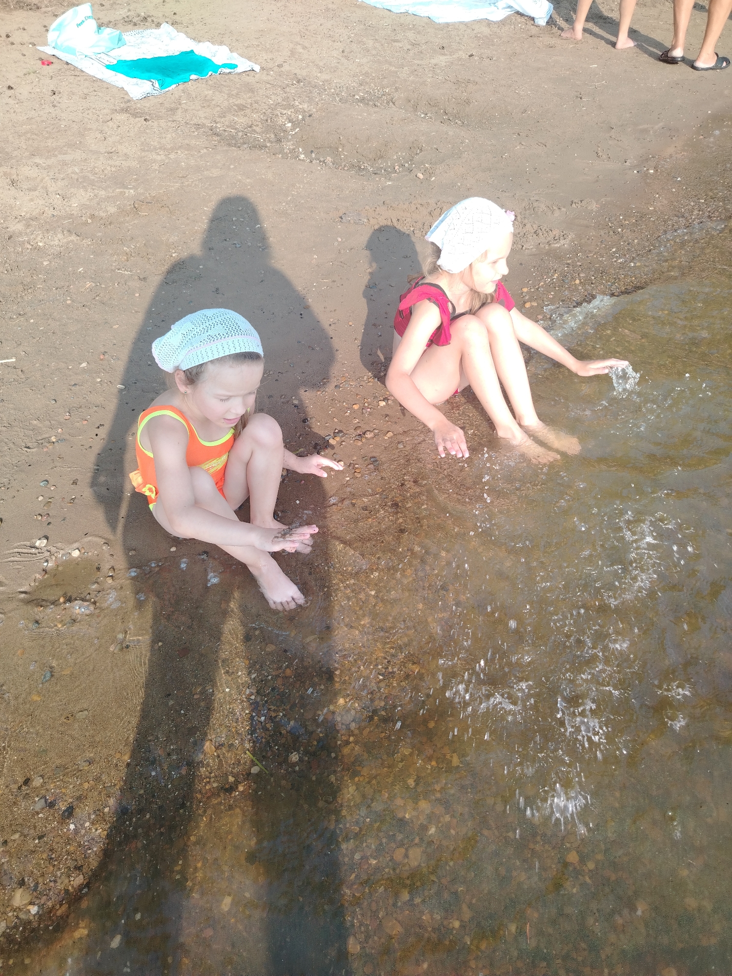 Two young girls are sitting on a sandy beach near the water's edge. The girl on the left is wearing an orange swimsuit and a white headscarf. She is looking down at her hands, which are in the shallow water. The girl on the right is wearing a red swimsuit and a white headscarf. She is reaching her hand into the water, creating splashes. Both girls appear to be enjoying themselves.
The scene is set on a sunny day, suggested by the shadows cast on the sand. The water is clear and shallow, with pebbles visible on the bottom. In the background, there is a light blue and white towel laid out on the sand, and the legs of at least two other people are visible, suggesting a casual outing at the beach. The overall atmosphere is one of relaxed summer fun. The location is Chaikovsky, Russia. There is no visible text in the image.