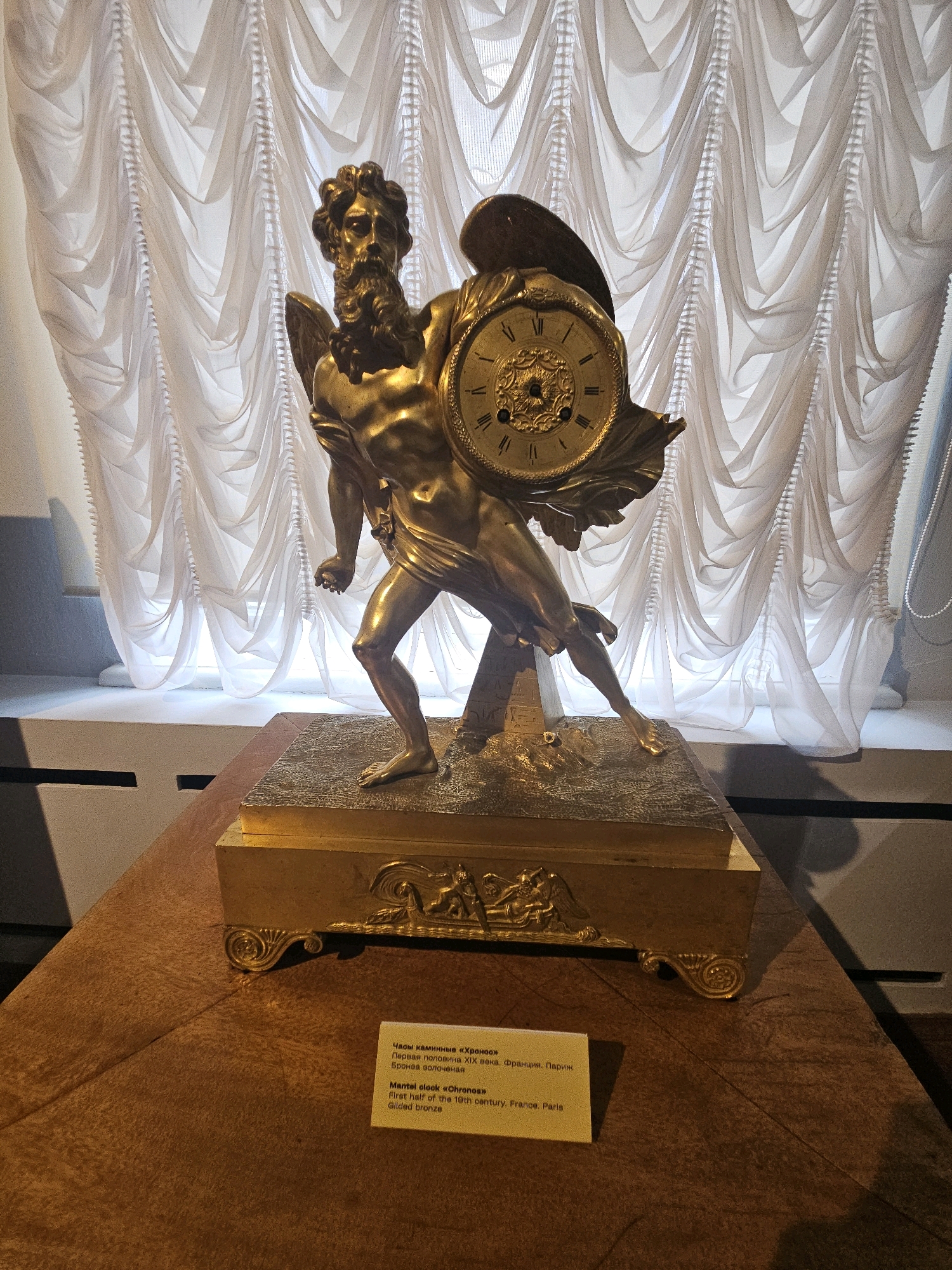 An ornate gilded bronze mantel clock, depicting a winged male figure with a beard holding the clock face, stands on a pedestal against a backdrop of draped white curtains. The clock is positioned on a wooden surface, possibly a table or display. A small yellow informational plaque is placed in front of the clock. The plaque contains text in both Russian and English. The Russian text reads: "Часы каминные «Хронос» Первая половина XIX века. Франция. Париж. Бронза золоченая". The English text reads: "Mantel clock «Chronos» First half of the 19th century. France. Paris. Gilded bronze". This indicates the clock is an artifact from the first half of the 19th century, originating from Paris, France, and is made of gilded bronze. The location is specified as the Ulyanovsk Regional Art Museum, an institution that exhibits various art forms and historical artifacts, including this mantel clock. The overall scene is well-lit, highlighting the detailed craftsmanship of the clock.