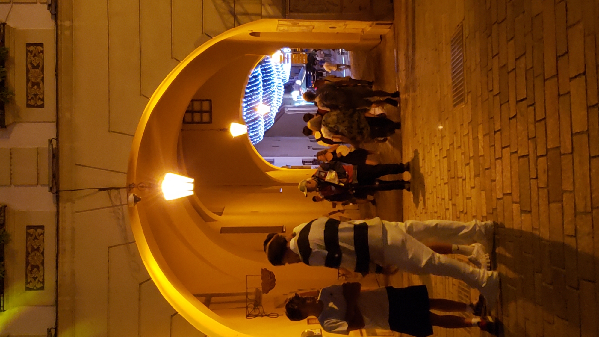 The image depicts a view through an illuminated arched passageway, likely in the historic city center of Prešov, Slovakia. The passageway is constructed of brick and is lit by warm yellow light from overhead lamps. A string of blue lights, possibly decorative, is visible in the distance, strung across the street beyond the arch. Several people are present, some standing in groups and others walking through the passageway. On the left, two young males stand facing each other, one wearing a white t-shirt and dark shorts, the other in a black and white striped shirt and white pants. Further in the passageway, a man in a cap and backpack is visible, as are other individuals in casual attire. The ground is paved with cobblestones. The architectural style of the buildings and the presence of decorative elements suggest a European urban setting.