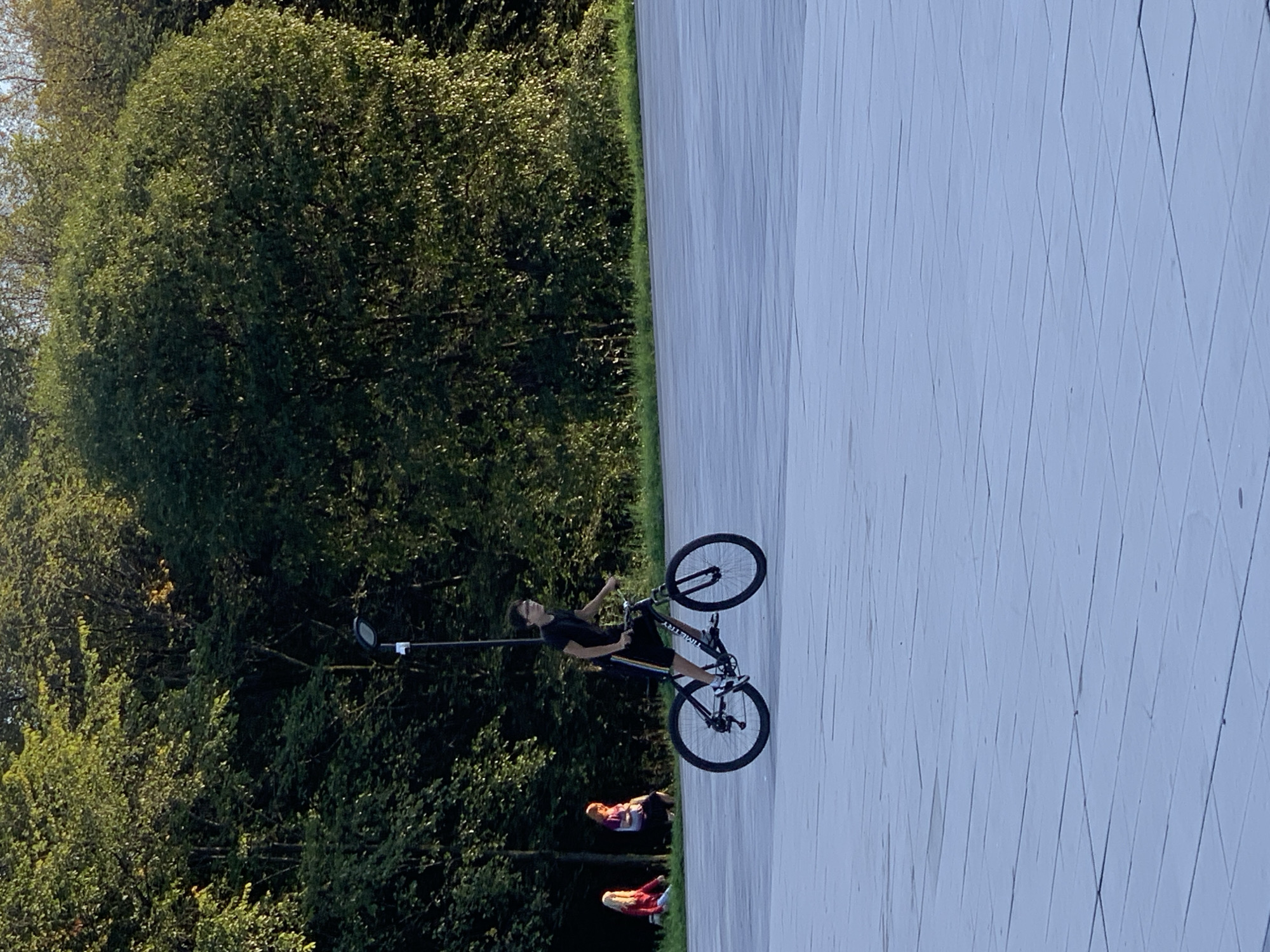 A young person with dark hair and glasses, wearing a dark t-shirt and shorts, rides a bicycle with black tires and a black frame on a paved area. They are positioned in the center-left of the frame. In the background, a line of dark green trees forms a dense backdrop. A lamppost is visible among the trees to the left of the cyclist. To the far left, two individuals are seated on the grass, with one wearing a red top and the other a purple top. The ground is a light-colored, tiled surface. The lighting suggests it is late afternoon or early evening, with sunlight casting shadows and illuminating the foliage. This scene is located in the city of Prokopyevsk, Russia.