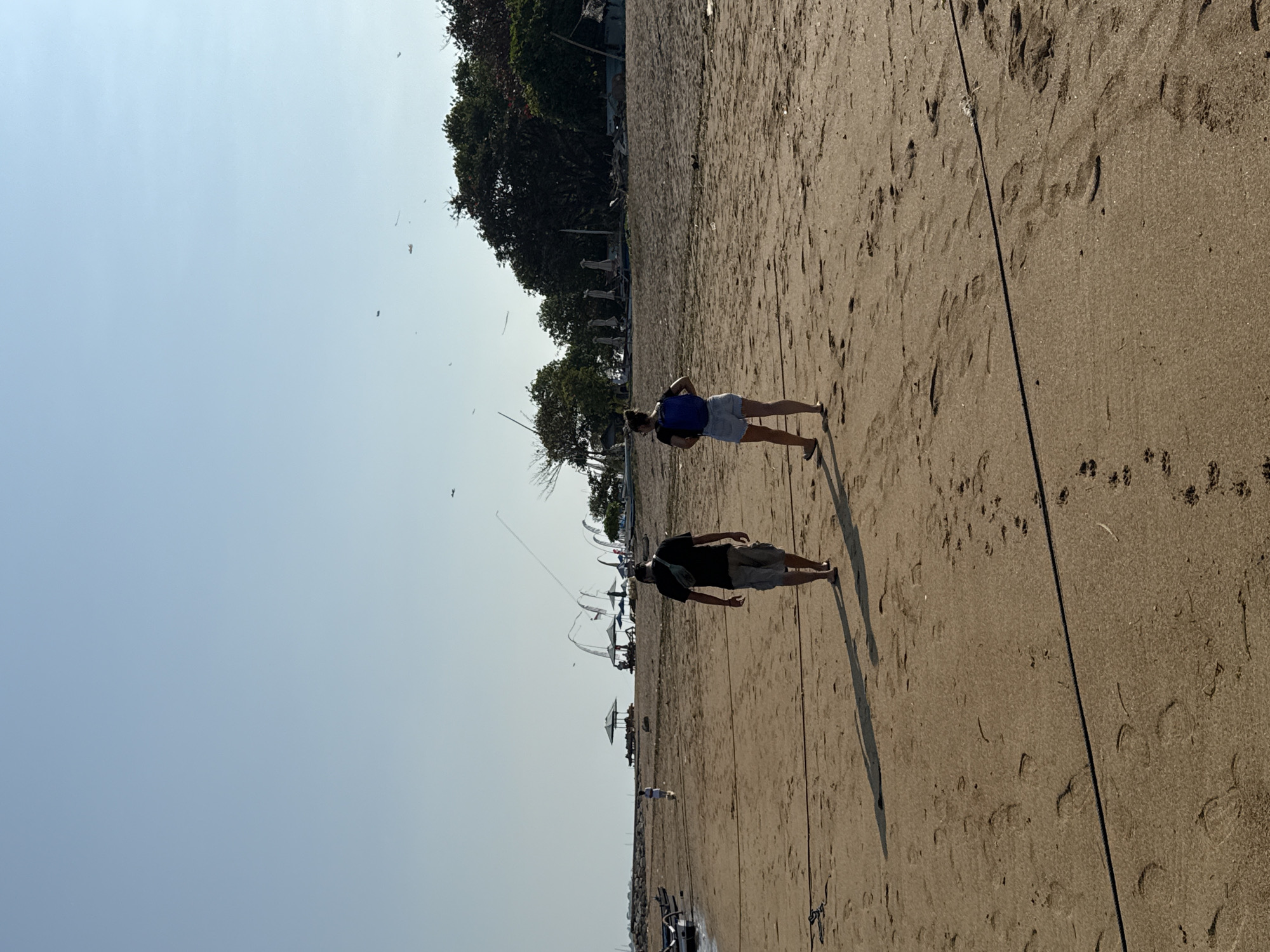 Two individuals are walking on a sandy beach in Denpasar, Indonesia, during the daytime. A male, facing away from the camera, wears a black t-shirt, grey shorts, and a fanny pack. A female, also facing away, is dressed in a dark blue top, denim shorts, and carries a blue backpack. They are walking towards the left side of the frame. The beach is populated with scattered footprints and a dark rope running horizontally across the sand. In the background, several beach umbrellas and structures are visible, along with trees and boats. The sky is clear and blue, with several distant kites visible. The overall scene depicts a relaxed day at the beach.