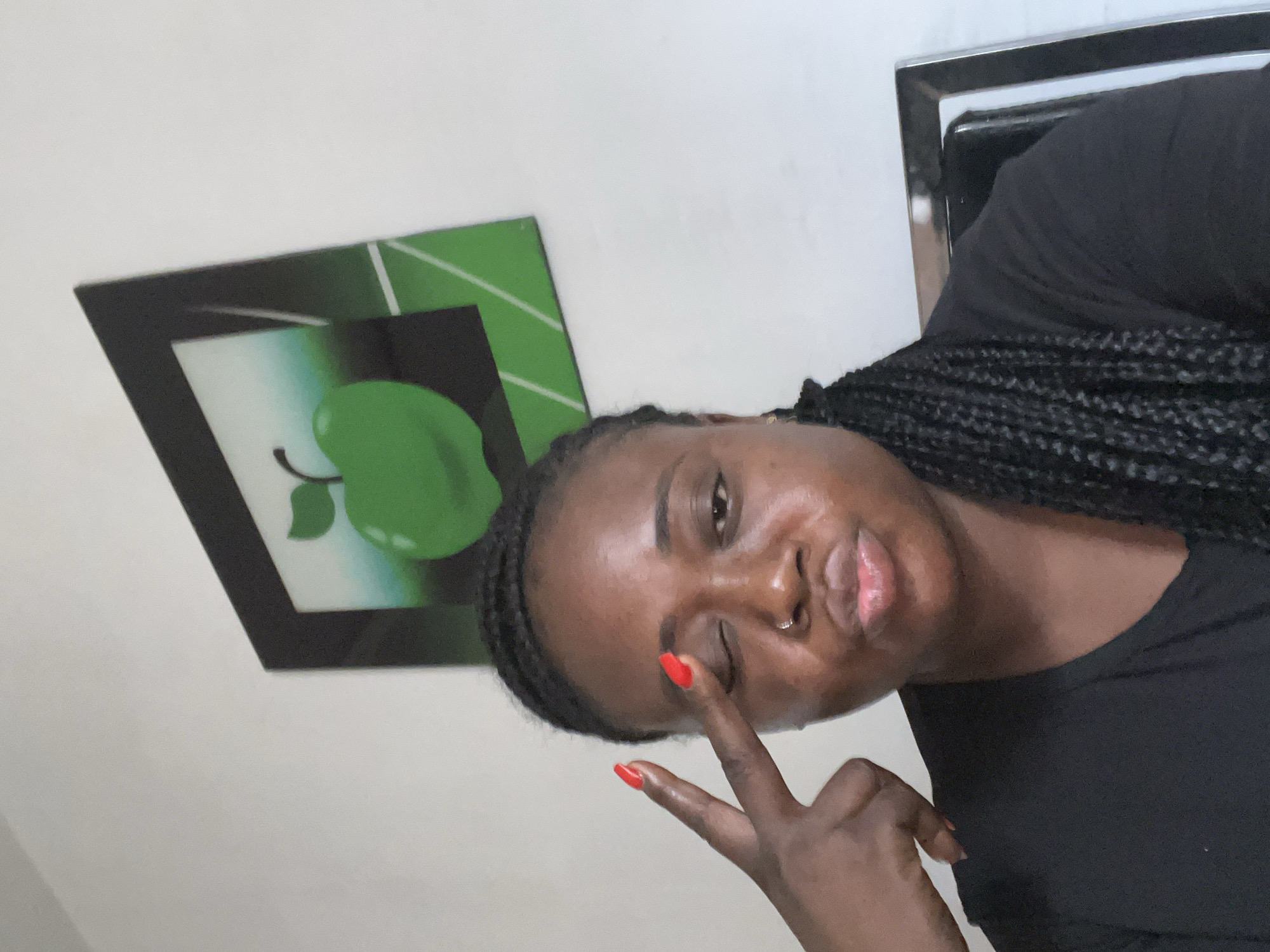 A close-up, angled portrait features a young woman in an indoor setting, taken in Jos, Nigeria. She has dark skin and her hair is styled in dark braids. She is wearing a plain black t-shirt. With a playful expression, she is seen winking with one eye while making a peace sign (V-sign) with her right hand, where her index and middle fingers are extended and adorned with bright red nail polish. A silver nose stud is visible on her right nostril.
Behind her, mounted on a light-colored wall, is a black-framed picture. The artwork within the frame prominently displays a stylized green apple with a leaf, set against a background of green and white, featuring geometric lines. To the far right of the image, a metallic object with black accents, possibly part of a cabinet or shelf, is partially visible. The lighting is soft and ambient, indicative of an interior space. No discernible text is present in the image.