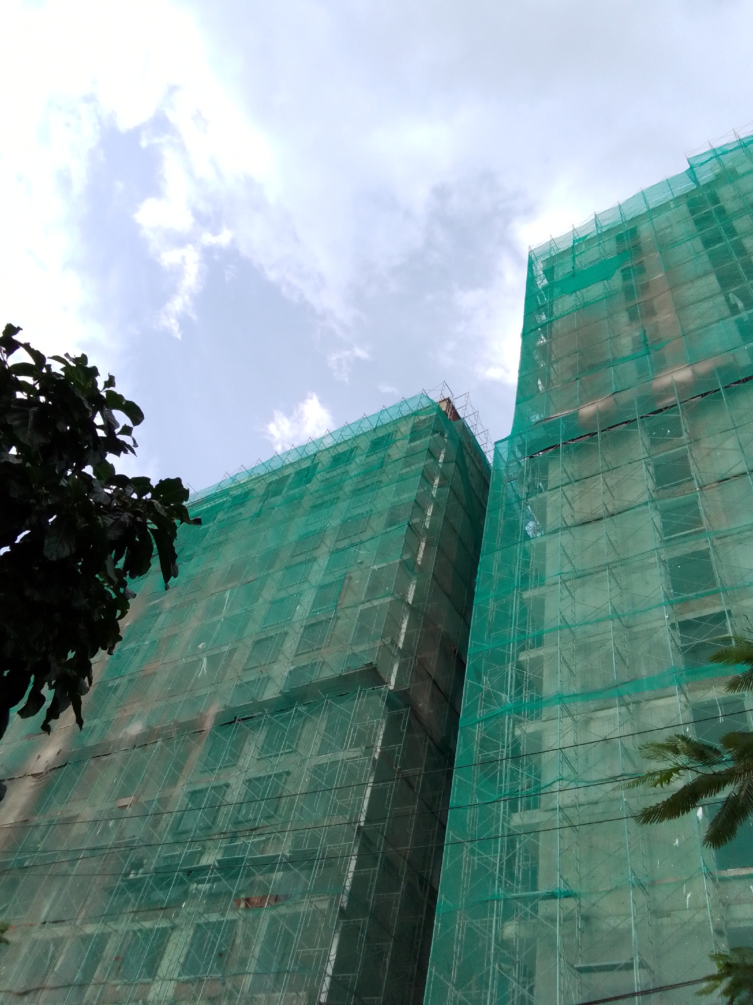 This is a low-angle, exterior, full shot of two under-construction high-rise buildings in Da Nang, Vietnam. The buildings are covered in green netting, a common sight during construction projects in the city. The netting is taut and covers the entire facade of both buildings, obscuring the details of their construction. The sky above is overcast with patches of white clouds. In the foreground, on the left side of the image, is a dark green tree with large leaves. On the right side of the image, a portion of a palm tree with fronds is visible. The overall impression is one of ongoing development and urban expansion.