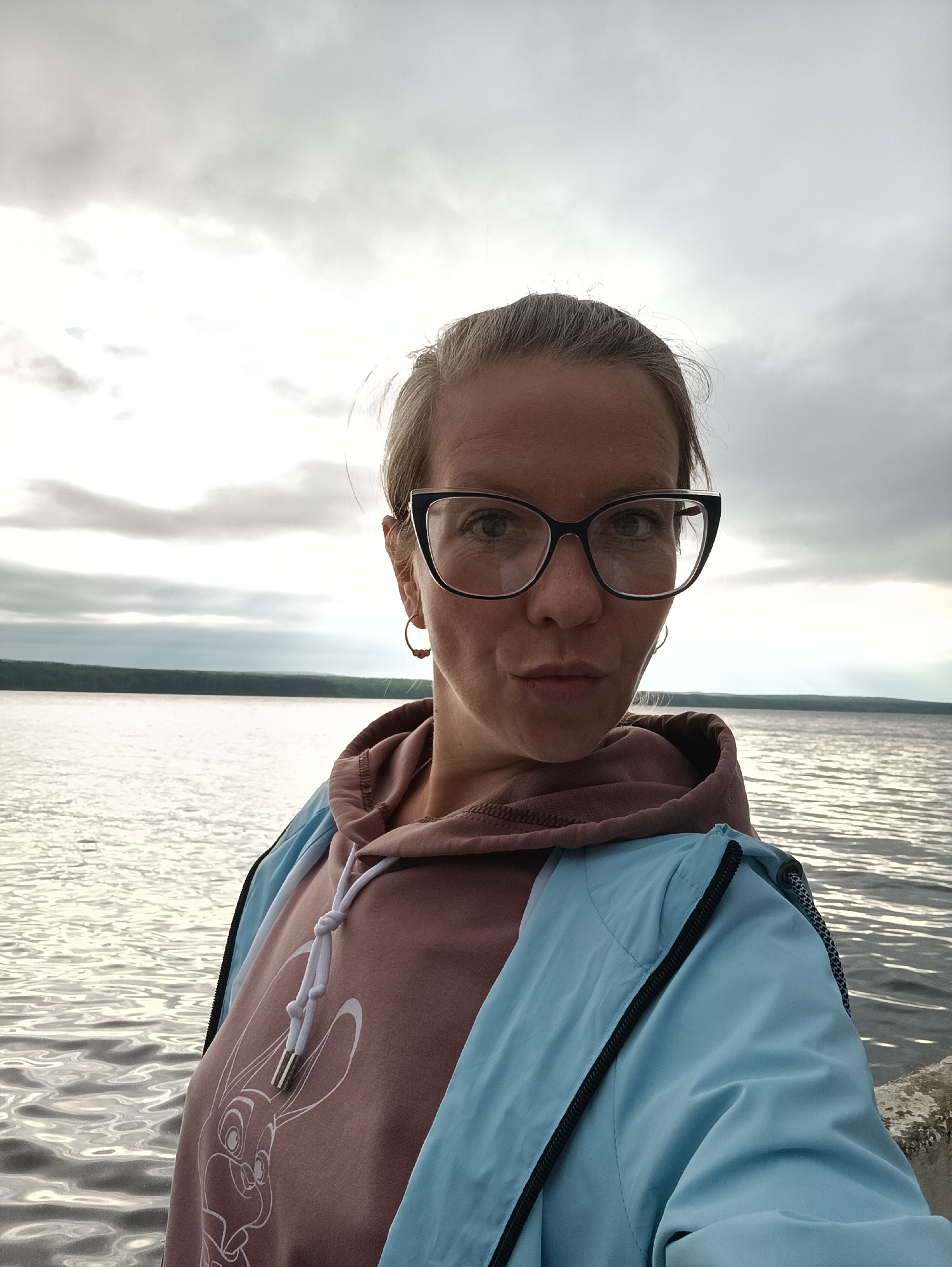 A woman with blonde hair pulled back and wearing black-rimmed glasses and hoop earrings is taking a selfie by a body of water. She is wearing a pink hoodie with a white rabbit design and a light blue jacket. The background shows a cloudy sky and a distant tree line across the water. The photograph appears to be taken outdoors, possibly at a pier or waterfront in the city of Chaikovsky, Russia, given the landscape.