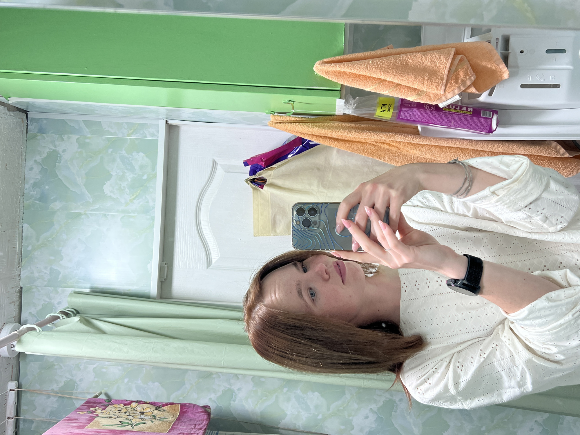 A young woman with shoulder-length reddish-brown hair and blue eyes takes a mirror selfie in what appears to be a bathroom or utility room. She is wearing a white, embroidered long-sleeved top and has a black smartwatch on her left wrist and silver bracelets on her right. She holds a smartphone, likely an iPhone, in a clear case with a topographic pattern. Her fingernails are painted a light pink/nude color.
The background, reflected in the mirror, shows a functional domestic space. Above her reflection is a vibrant green painted shelf or cabinet. Below it, folded orange towels are visible, alongside a white plastic shelf or part of an appliance. A purple package, possibly a hygiene or cosmetic product, is resting among the towels. A small yellow label on one of the towels shows Cyrillic text, including "ЛАП 70ВК". The wall is covered with light green and white marble-patterned tiles. A white door with a curved decorative panel is also reflected. To the left, a light green rolled-up shower curtain or fabric is hanging, and below it, a small pink item with a floral pattern, possibly a mat, is placed on a clothes drying rack. The Cyrillic text on the purple package appears reversed, showing "РЕЛАКС" (RELAX) and words like "НЕЖНЫЙ" (NEZHNYI - tender) and "АПЕЛЬСИНОВЫЙ АРОМАТ" (APELSINOVYI AROMAT - orange aroma). The woman looks directly into the camera with a neutral expression. The scene suggests an indoor setting, likely during the daytime.