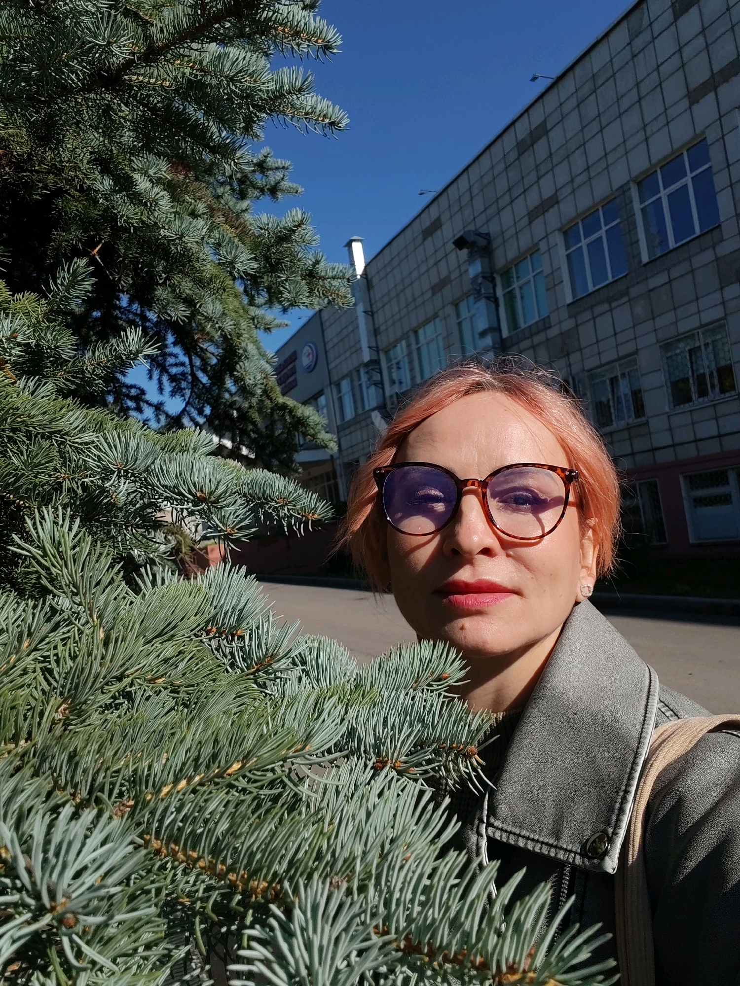 The image shows a woman with short, pinkish hair and glasses, posing for a selfie outdoors. She is wearing a gray jacket. In the foreground, there is a pine tree branch with blue-green needles. Behind her, a modern building with a grid-like facade and numerous windows is visible under a clear blue sky. The scene appears to be set in an urban environment, possibly a public space or a courtyard. The location is identified as Chaikovsky, Russia.