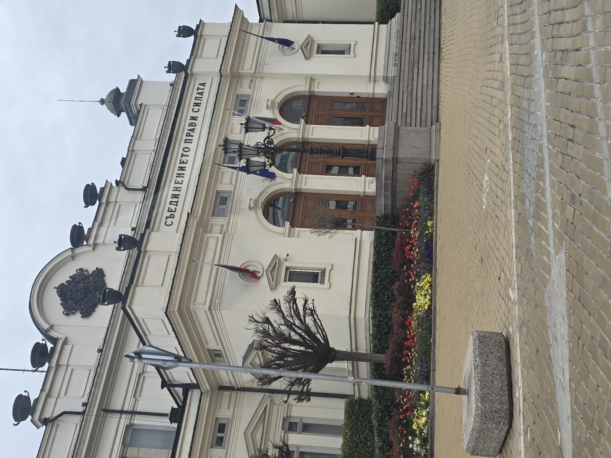 The image captures a low-angle shot of the National Assembly of Bulgaria building in Sofia. The building is white with dark brown doors and windows. The facade displays the inscription "Съединението прави силата" (Unity Makes Strength) in Cyrillic. Bulgarian and EU flags hang from the building. Below, a flowerbed is full of colorful flowers. A bare tree and a street light pole are visible in the foreground. The street is paved with light brown bricks. The sky is overcast. A stone block is on the right side of the sidewalk. No people are visible.