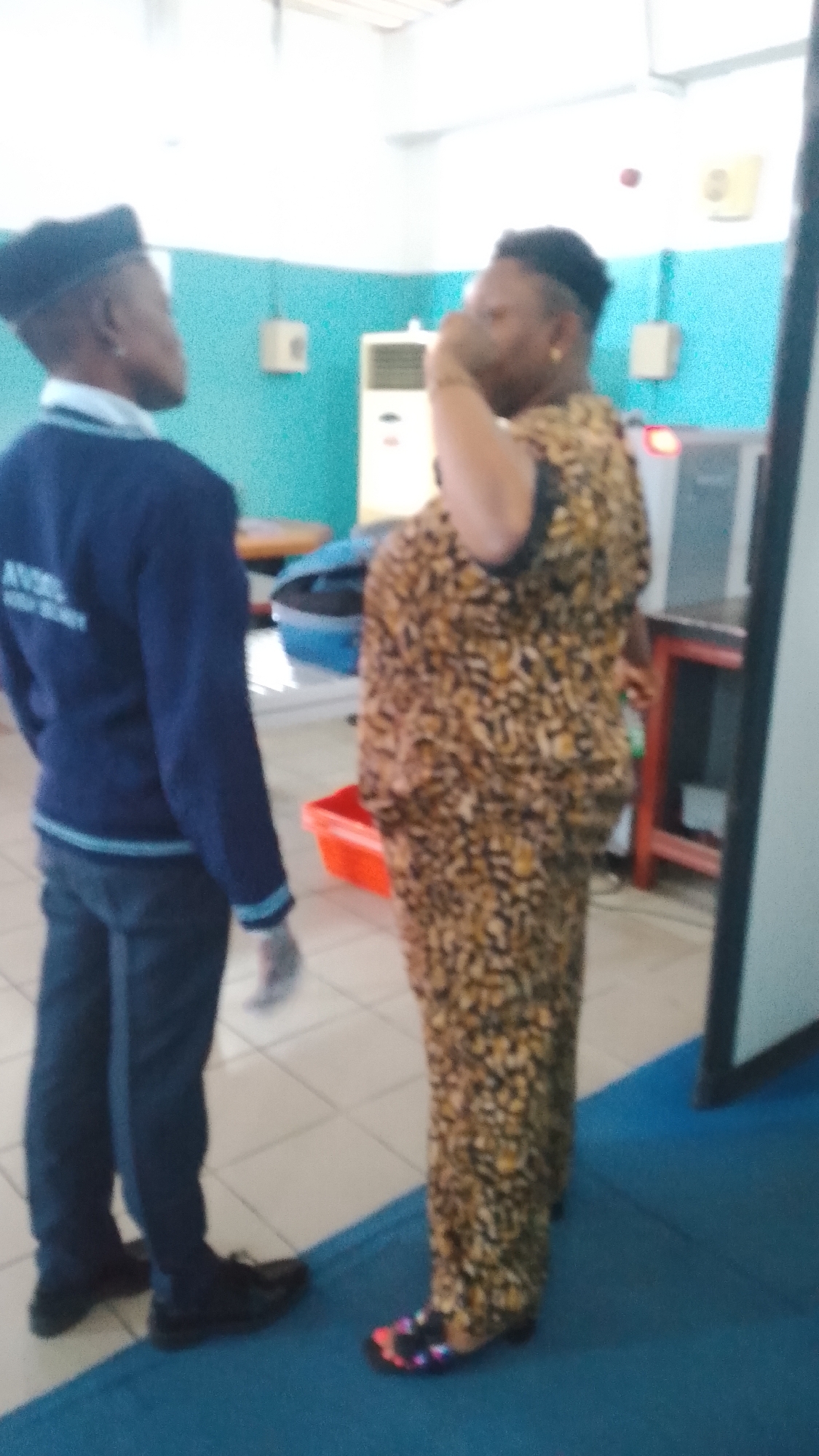 Two individuals are standing in what appears to be an indoor space, possibly an office or school setting in Orerokpe, Nigeria. The person on the left, wearing a navy blue sweater and dark trousers, has their back to the camera and is looking to the right. A glimpse of lettering is visible on the sweater. The person on the right, wearing a brown and black patterned dress and colorful sandals, is facing the person on the left and has their right hand raised to their face, appearing to be speaking or adjusting something. The environment includes a light-colored tiled floor, turquoise walls, and various objects in the background, such as a white air conditioning unit, what looks like a desk or table, and some shelving with items on it. The lighting is bright, suggesting an overhead light source.