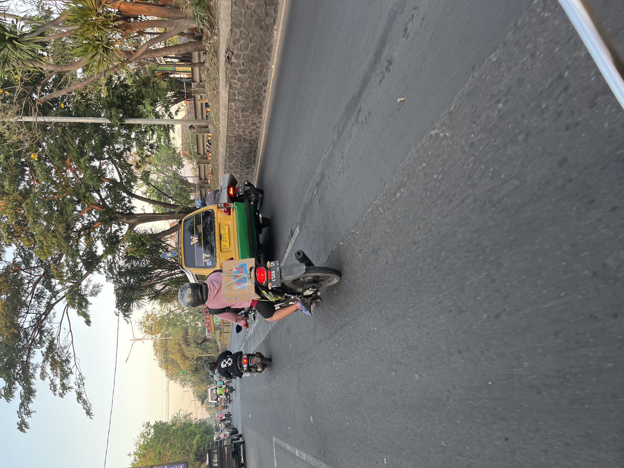 An over-the-shoulder, slightly blurred, eye-level shot depicts a street scene in Ngaban, Indonesia. The foreground is dominated by a motorcycle and its rider, viewed from behind and slightly to the side. The rider wears a black helmet and a pink hoodie, with a box strapped to the back of the motorcycle. The motorcycle itself is black with a license plate reading "L 4508 SF". In the middle ground, a yellow and green public transport van is visible, with a license plate indicating "H 1002 UR". Other motorcycles and a truck are also present on the road, indicating moderate traffic. The environment features a paved road, a stone wall on the right side of the road, and trees and vegetation lining the street. The sky is clear with a warm, orange hue, suggesting either sunrise or sunset.
