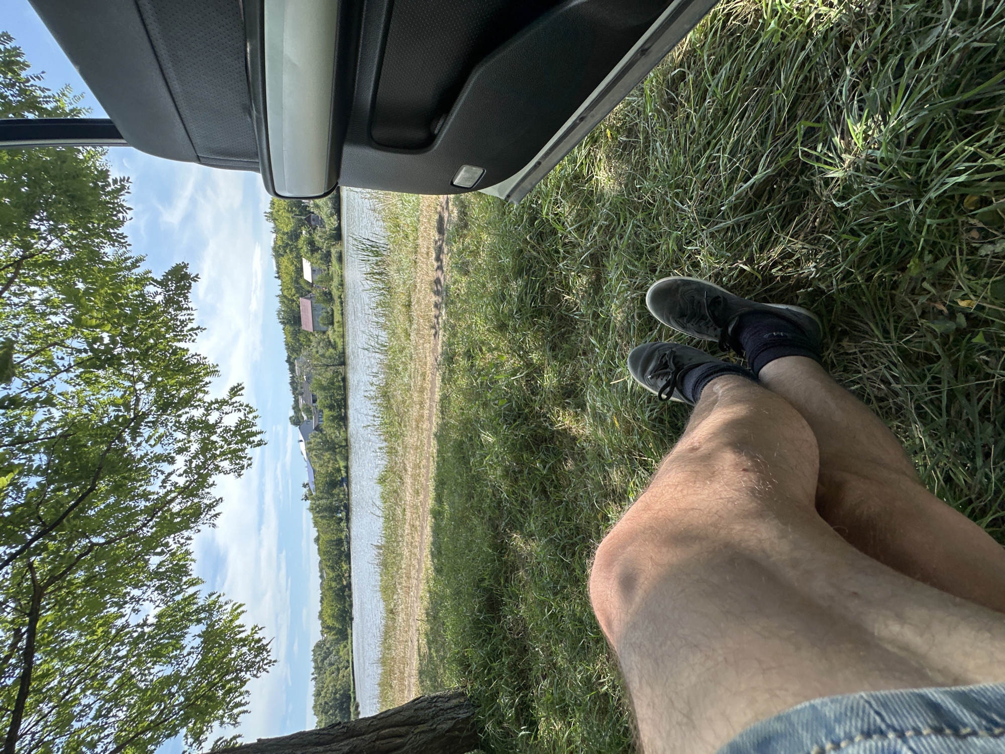 A person's legs are visible in the foreground, crossed at the ankles, wearing dark sneakers and dark socks. They are sitting on a grassy area near a body of water, possibly a lake or river. To the right, part of a car door is visible, with its interior and exterior trim in view. In the background, across the water, is a shoreline with trees and several houses with red and white roofs, suggesting a rural or suburban setting. The sky is blue with scattered clouds. The scene appears to be in Makiv, Ukraine, as indicated.