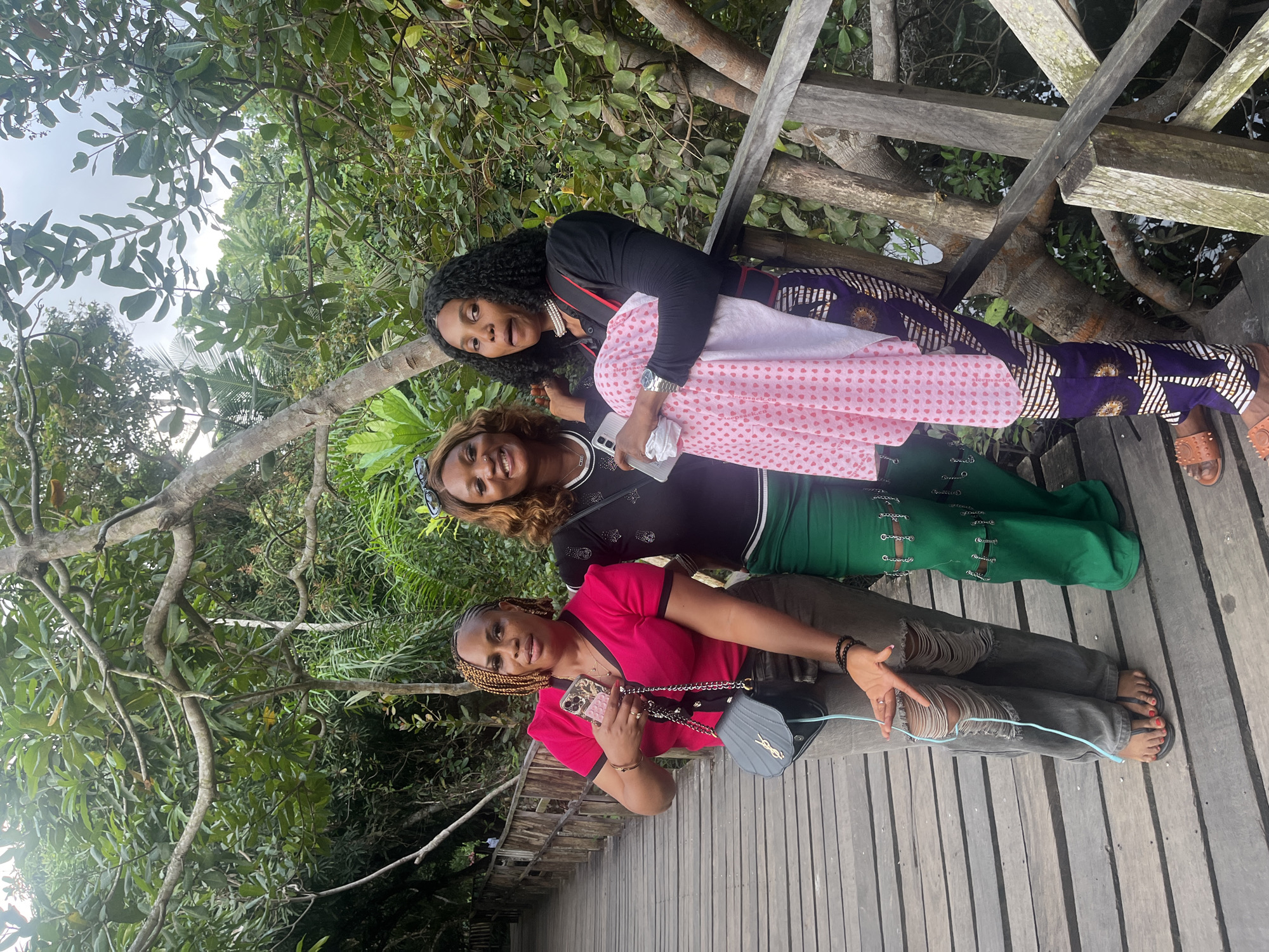 Three women stand on a wooden boardwalk surrounded by lush vegetation. The woman on the left wears a pink top with black trim and ripped gray jeans. She holds a phone and a black purse. The woman in the middle has wavy brown hair and wears a black top with a green skirt. The woman on the right has curly dark hair and wears a black long-sleeved top and patterned pants. She holds a pink and white patterned cloth. The boardwalk appears to be in a natural setting, possibly a park or a nature reserve within Lekki, Nigeria. The trees are dense and green, with sunlight filtering through the leaves.