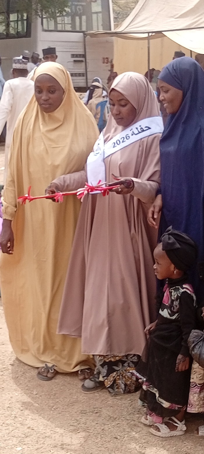 An outdoor, eye-level photograph captures a daylight scene in Kano, Nigeria, featuring four individuals in the foreground and a larger gathering in the background. In the left foreground, a woman wearing a mustard yellow hijab and full-length abaya holds the left end of a thin brown stick adorned with red and white ribbons. To her immediate right, a woman in a light brown hijab and abaya wears a white sash bearing Arabic text "حفلة 2026" (Ceremony 2026); she holds the right end of the same ribbon-decorated stick with both hands, her right hand displaying henna patterns. Adjacent to her on the right, a woman in a dark blue hijab and abaya stands partially visible. To the far right, a young child, dressed in a black long-sleeved top with colorful patterns, a black skirt, and a black headwrap tied in a bow, is positioned, looking left. The ground is unpaved and appears dusty. The background includes blurred figures, a light-colored building with dark-framed windows, a white tent structure, and a wall section marked "13".