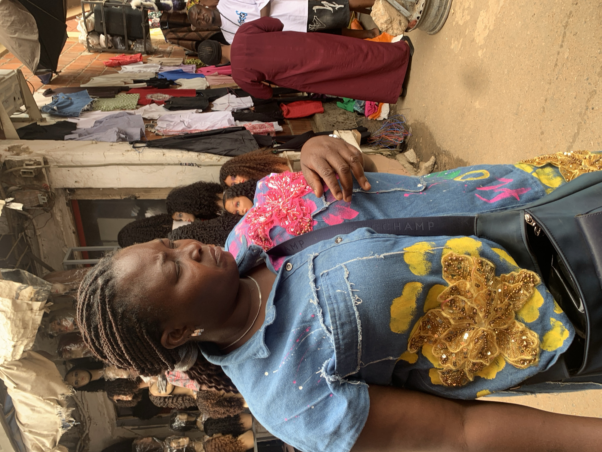 A woman with braided hair is lying down, her eyes closed, wearing a blue denim shirt adorned with colorful paint splatters and large floral appliques. One applique is a bright pink flower with sequins, and another is a golden sequined flower. She has a silver necklace and earrings. In the background, several other people are visible, including a man in a white t-shirt and black shorts and another person wearing a burgundy robe. Various items are spread out on the ground and on shelves, including clothing, what appear to be wigs or hair extensions, bags, and electrical wires. The scene appears to be a marketplace, possibly an outdoor stall or shop, with a ground paved with bricks and dirt. The overall impression is one of a bustling, vibrant, and somewhat chaotic environment, potentially in Abuja, Nigeria, given the location context. The lighting suggests it is daytime.