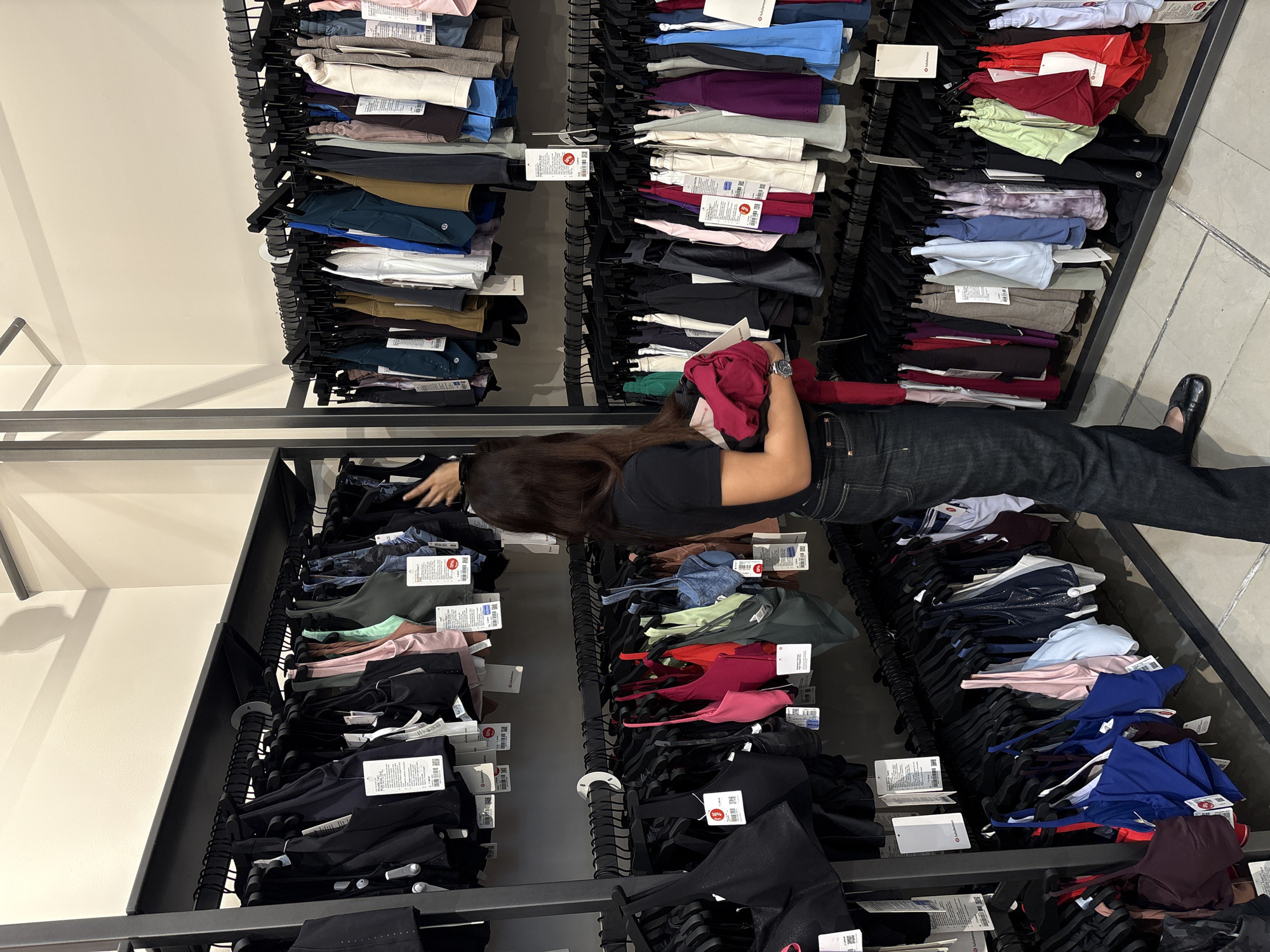 This indoor photograph depicts a retail store environment, likely an athletic wear shop, potentially Lululemon, situated in San Francisco Ocotlán, Mexico. The main subject is a woman with long, dark hair, wearing a dark t-shirt and dark jeans, actively browsing items on a clothing rack. She is seen reaching into a rack, with a folded red garment held in her left arm, and her right hand sifting through other clothing. A silver wristwatch is visible on her left wrist.
The scene is dominated by multiple black metal clothing racks, densely packed with neatly folded athletic wear, including leggings, sports bras, and tops. These garments are displayed in a wide spectrum of colors such as black, various shades of blue, red, green, purple, grey, white, beige, and pink. Each item is attached to a black hanger, and many feature white price tags. Visible text on these tags includes the "lululemon" brand name and logo, along with some red "50% OFF" indicators, suggesting a sale. The store's floor is made of light-colored tiles, and a plain white wall serves as the background, illuminated by bright indoor lighting. The atmosphere suggests a typical daytime shopping experience.