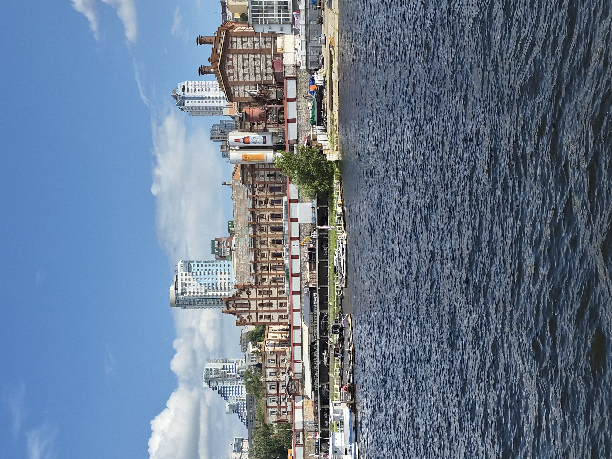This is a wide shot of the Samara embankment in Samara, Russia, on a clear, sunny day with some clouds. The image shows a body of dark blue water in the foreground, with small waves. In the middle ground, there is a row of moored boats and a pier, with several small vessels docked. Behind the pier is the Samara embankment, featuring a variety of buildings.
The most prominent structures are industrial buildings, some appearing to be old factories with red brick facades and large windows. One building has large cylindrical tanks on its side, one of which is painted with a picture of a beer mug. There are also modern high-rise buildings in the background, some with glass facades. The embankment area shows parked cars and some construction or storage materials. The sky is a bright blue with fluffy white clouds. The scene depicts a waterfront urban environment with a mix of historical industrial architecture and modern development.