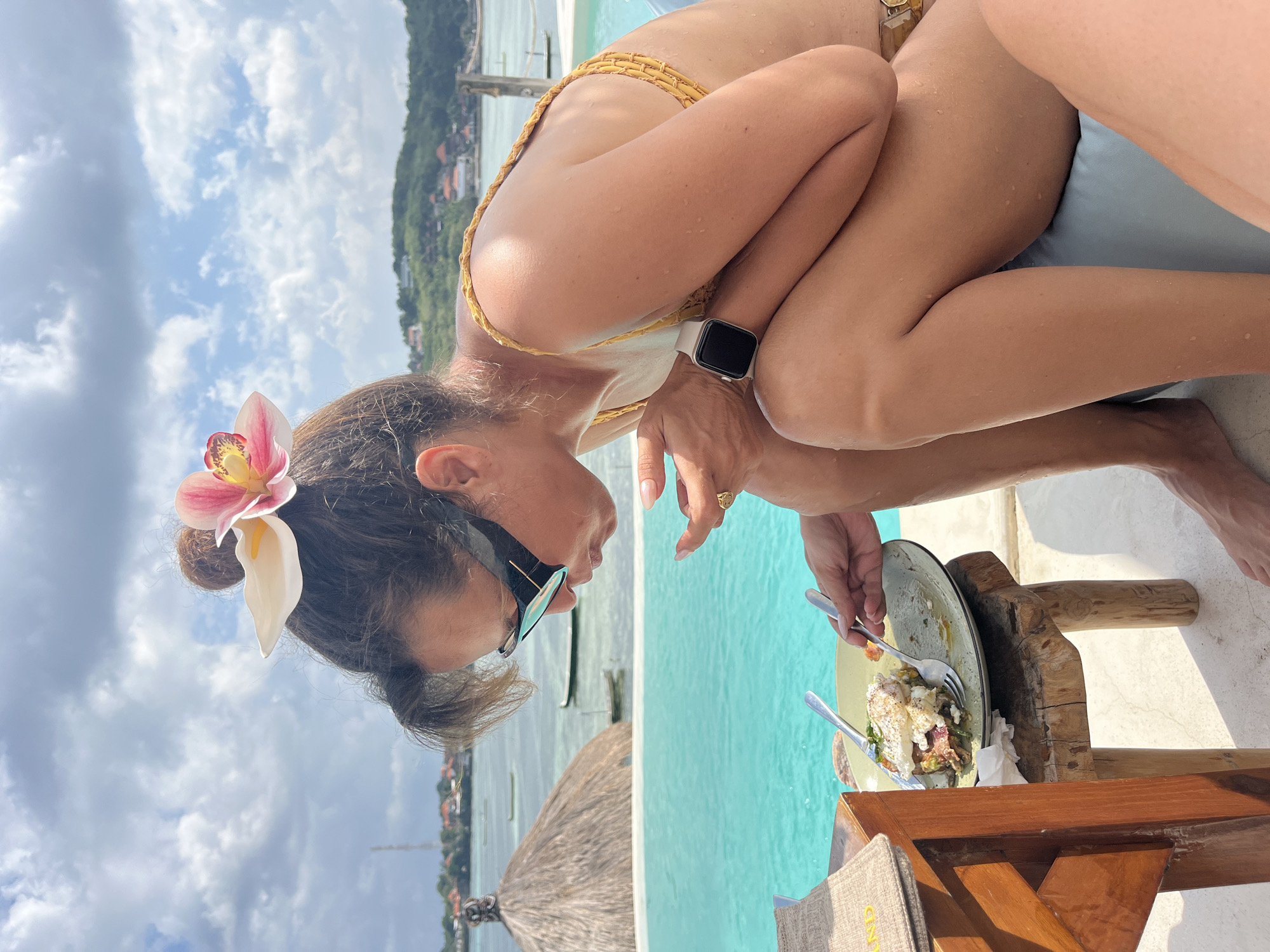 A woman with a flower in her hair is eating a meal next to an infinity pool in Nusa Penida, Indonesia. She is wearing sunglasses and a yellow bikini. The pool overlooks the ocean, with boats and buildings visible in the background. A thatched-roof hut is also visible near the pool. The woman is holding a fork and knife, and a plate of food is on a wooden stool beside her.