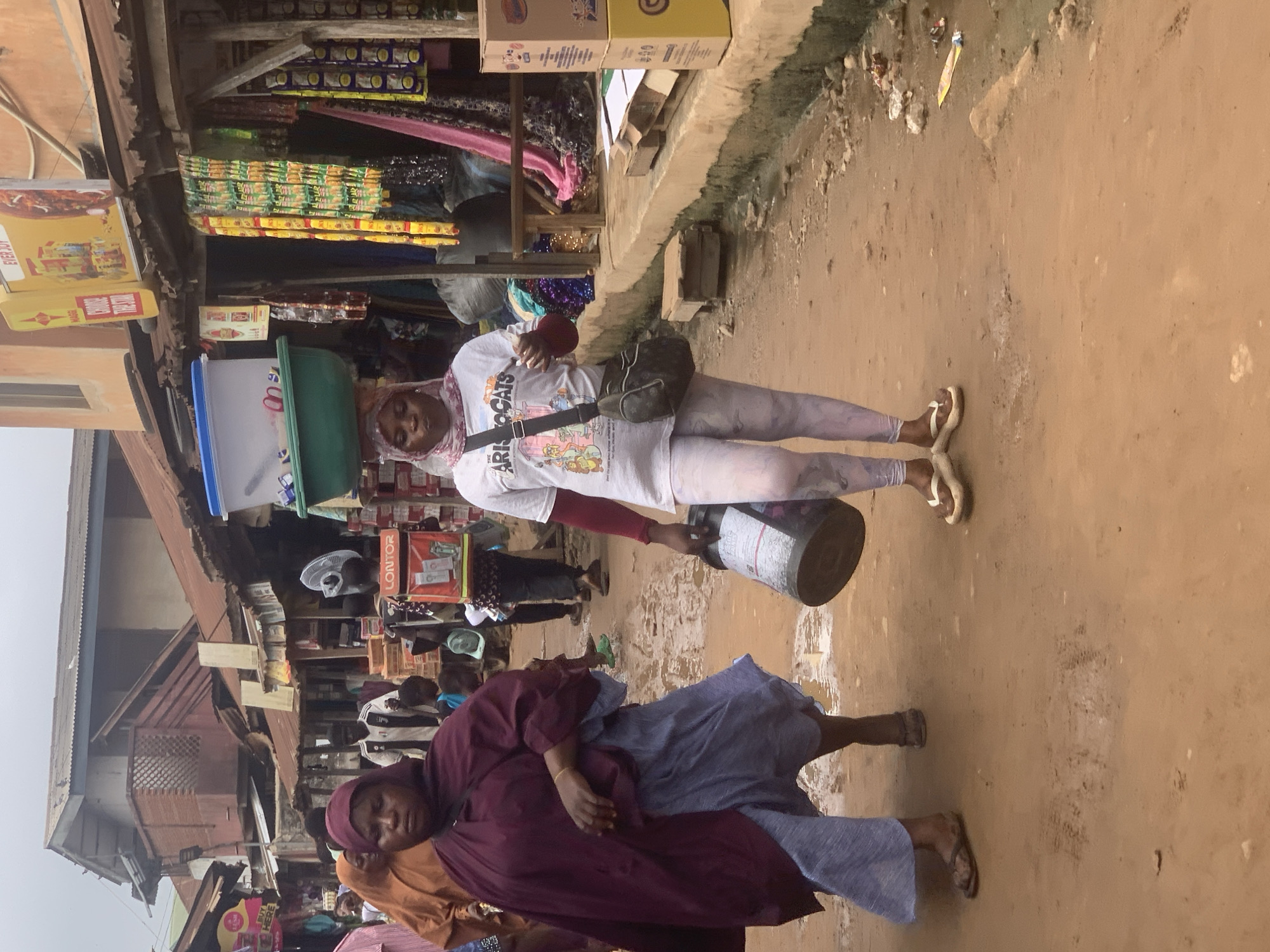 The image depicts a street scene in Shagamu, Nigeria.
A woman in a white t-shirt with an image of The Aristocats, light-colored pants, and sandals is walking on the street. She has a patterned headscarf and a black bag strapped across her chest. She is carrying two large containers; one stacked on her head and the other in her right hand.
Another woman is also seen walking on the street in front of her. She is wearing a maroon top and a blue skirt. She also has a patterned headscarf and sandals.
There are several shops visible in the background, with various goods for sale. Other people are visible in the background as well. The ground appears to be dirt or packed earth.