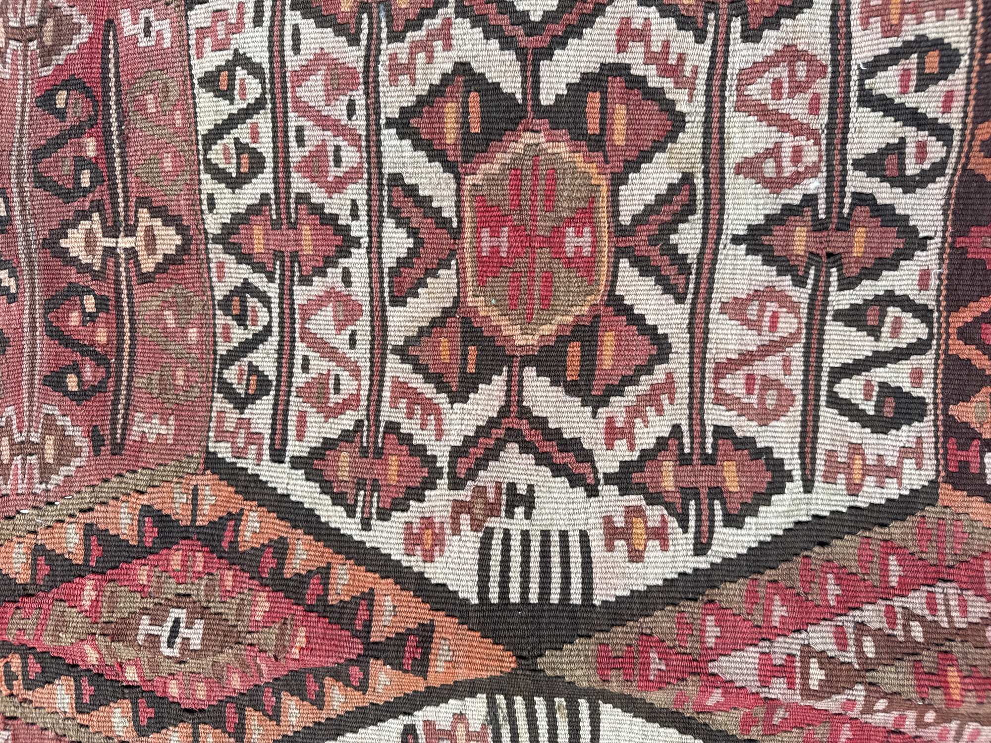 The image is a close-up, overhead shot of a colorful woven textile, likely a rug or tapestry. The pattern is intricate and geometric, featuring diamond shapes, zigzags, and stylized figures in shades of red, pink, brown, black, white, and orange. The textile appears to be made of natural fibers, with visible threads and a slightly uneven texture. The lighting is even, and there are no shadows, suggesting it was photographed in a well-lit indoor environment. There are no people, actions, or interactions visible. The provided location, Dilijan, Armenia, suggests the textile might be of Armenian origin or found within a context related to Armenian culture.