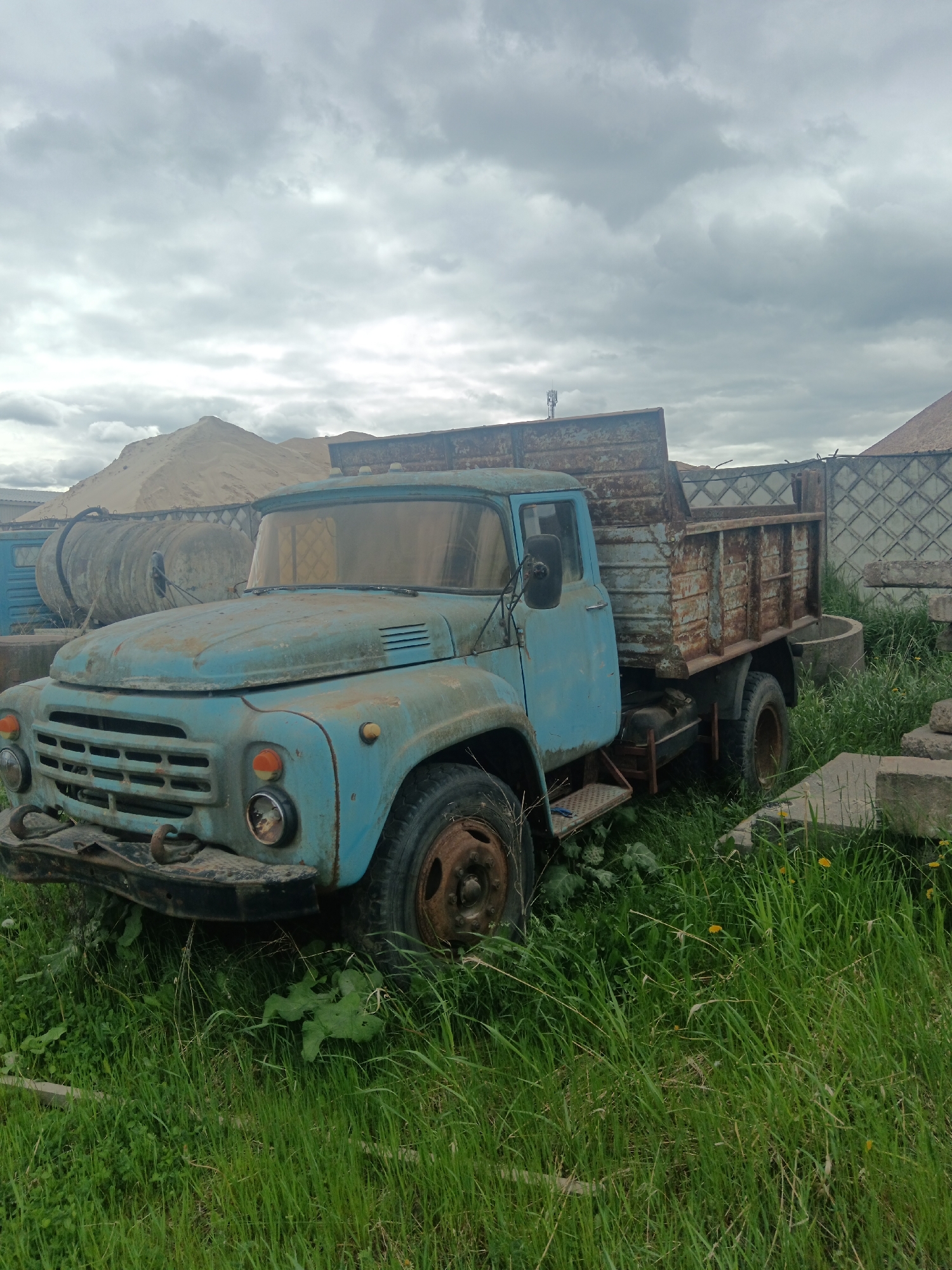 The image depicts an old, dilapidated blue truck parked in a grassy area, presumably in Яльгелево, Russia. The truck has a rusty bed and is surrounded by tall grass and weeds. In the background, there is a pile of sand, a large, cylindrical tank, and a concrete fence. The sky is overcast. No people are visible.