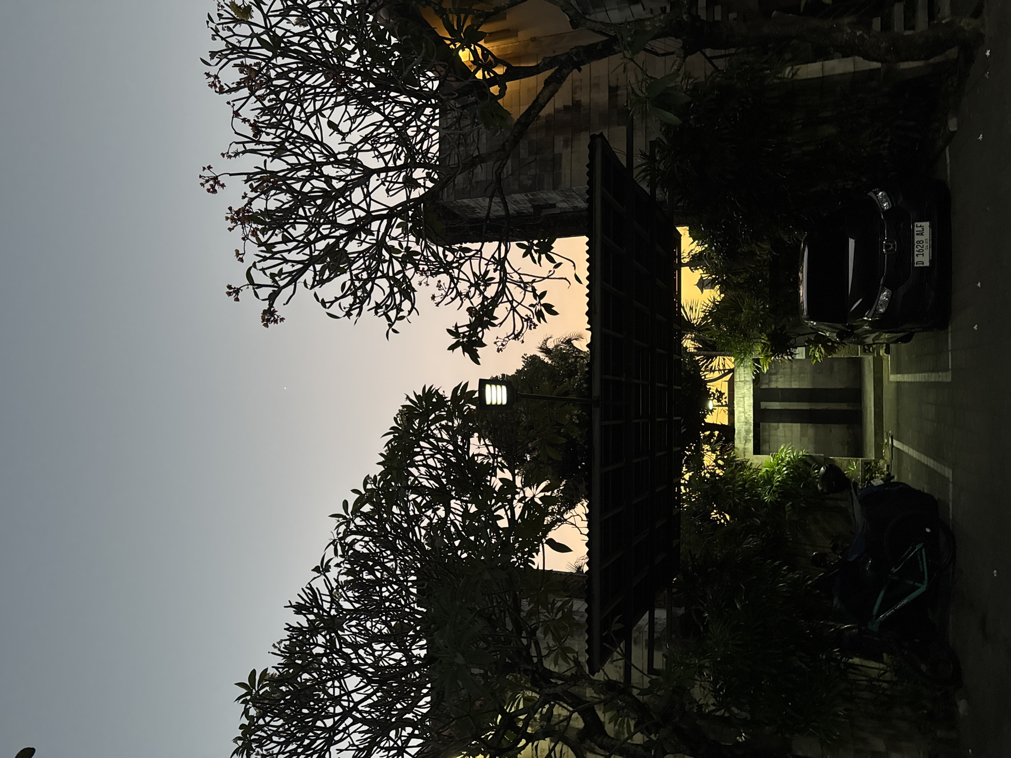 At dusk in Denpasar, Indonesia, a paved driveway is flanked by dark green foliage and two silhouetted trees with bare branches. A black car with license plate D 1628 ALF is parked in the driveway, alongside a motorcycle covered with a dark tarp. Beyond the car, a double door is visible set into a stone wall. A covered walkway with a corrugated metal roof extends over the driveway, supported by columns and adorned with a single modern lamp. The sky above displays a gradient of pale grey and soft orange, with a faint planetary object visible in the upper portion.