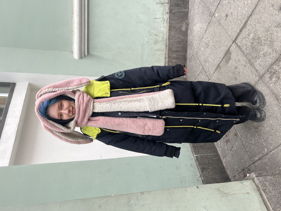 A single individual, appearing young, stands centered on a grey concrete sidewalk, facing the camera. The person has blue-dyed hair, pale skin, light-colored eyes, and multiple facial piercings including eyebrow studs, nostril studs, and lower lip rings. They wear a long, dark navy or black puffer coat with bright yellow lining visible at the collar and yellow drawstring cords with black stoppers. A large, fluffy pink and off-white hood featuring long, rabbit-like ears is worn, with an integrated wide scarf draping down the front. The scarf is pink on one side and off-white on the other. Dark pants and black, chunky-soled boots complete the attire. The individual exhibits a slight smile with the tongue lightly exposed. The background consists of a building wall painted a light pastel green, featuring a subtle horizontal molding line. A window frame or ledge is partially visible in the upper left. The concrete sidewalk displays visible cracks and minor debris. The scene is illuminated by diffused natural light. The specific location is a street in the city of Unknown, Unknown.