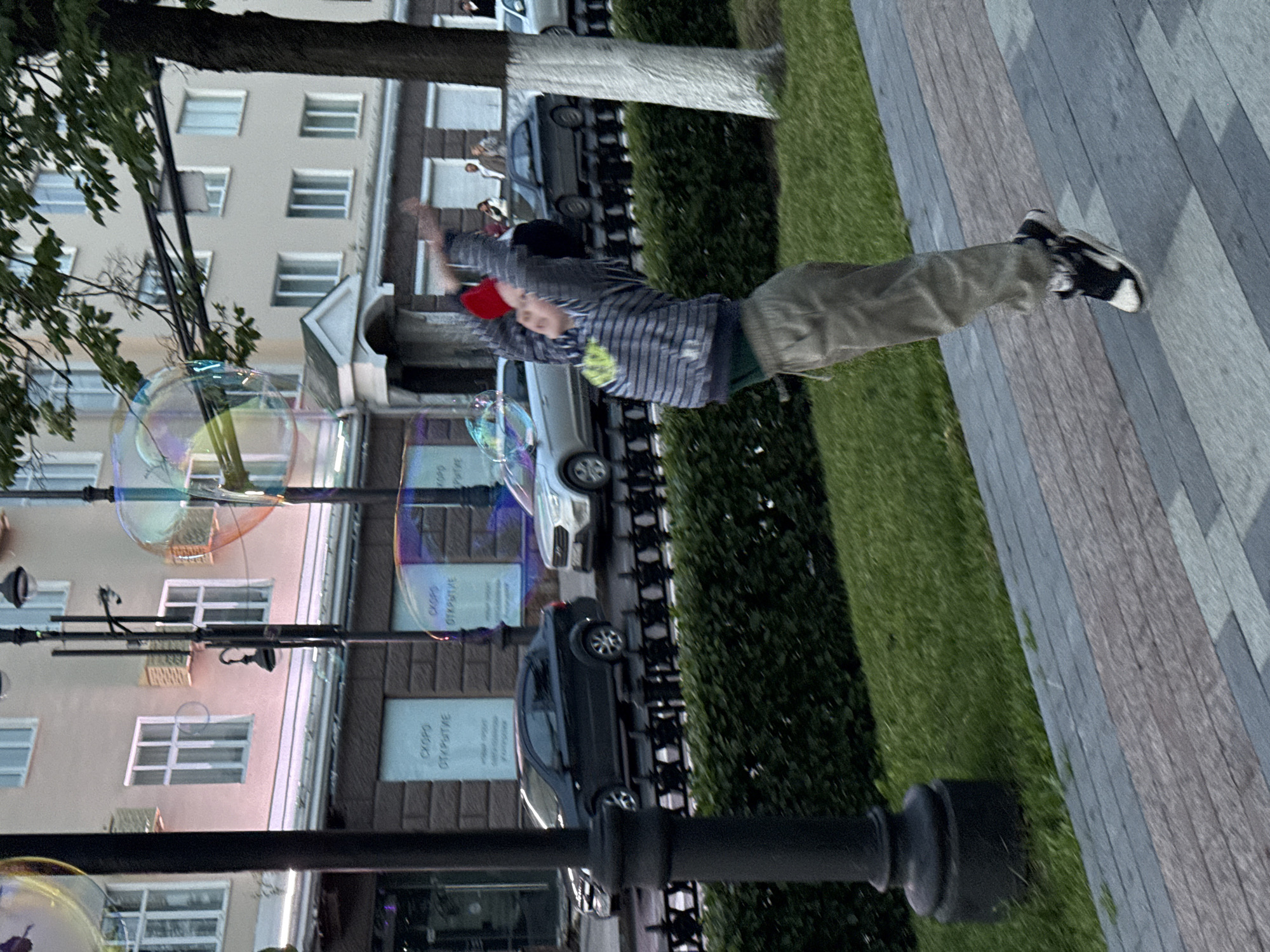 The image captures a young person in mid-air, appearing to jump or playfully pose, on a sidewalk paved with rectangular blocks. The person is wearing a striped long-sleeved shirt, khaki pants, and black and white sneakers. A red cap is on their head, and their arms are outstretched. Large, iridescent soap bubbles float in the background, some catching the light.
Behind the sidewalk and the person is a green lawn bordered by a black metal fence. Beyond the fence, a street is visible with several cars parked and driving, including a black sedan and a silver hatchback. The background features buildings with multiple windows and what appear to be advertisements on their facades, one reading "СКОРО ОТКРЫТИЕ" (Soon Opening). The architecture and the signage suggest an urban environment in Russia.
Based on the visual cues, such as the architecture of the buildings and the style of the advertisements, this scene is likely located in the city of Perm, Russia. Specifically, the appearance of the buildings and the streetscape are consistent with urban areas in Perm.