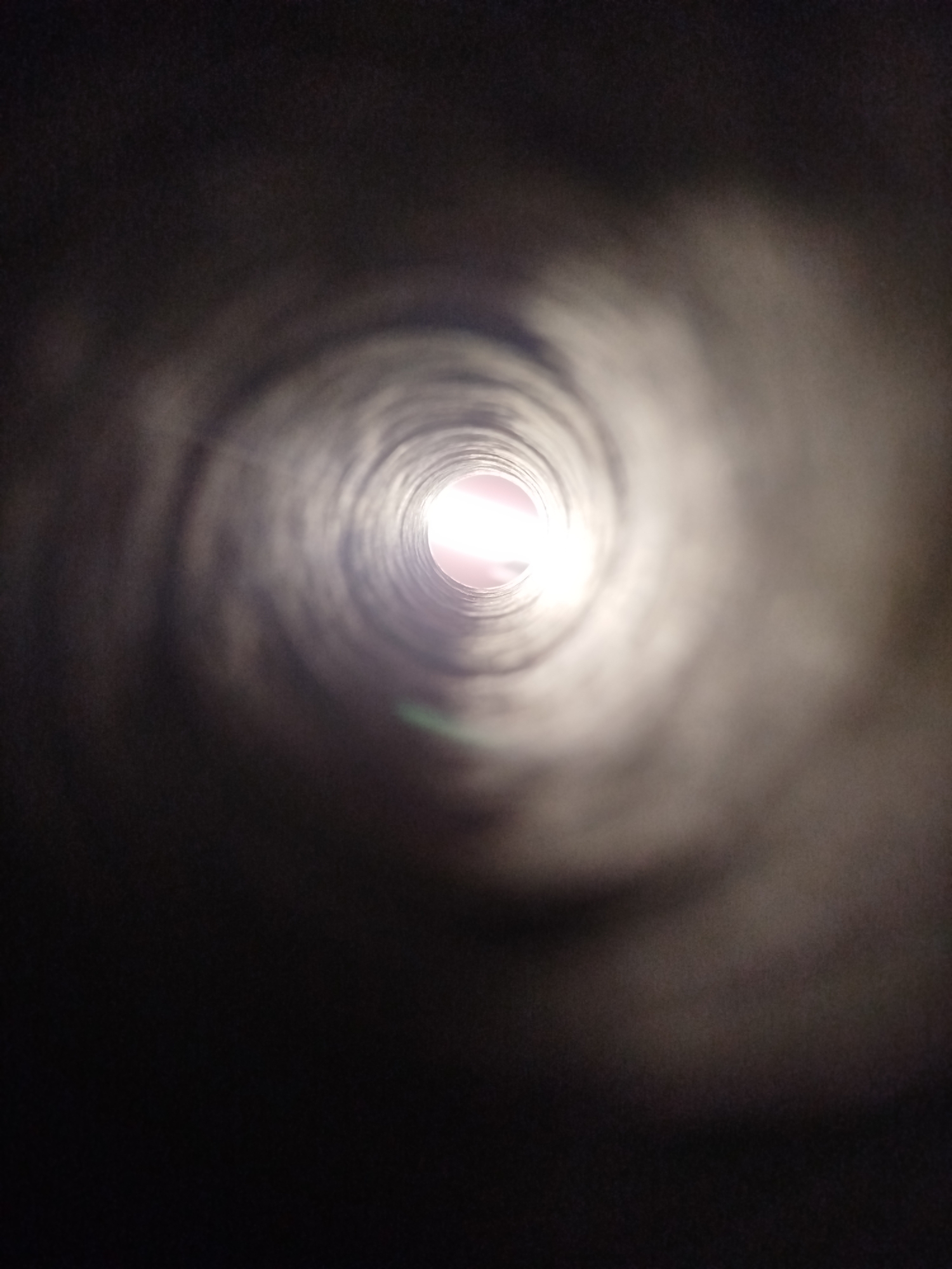 The image shows a view from inside a cylindrical, dark tunnel looking outwards towards a bright light source. The tunnel walls are textured and appear to be made of a rough material. The light at the end of the tunnel has a pinkish hue and appears to be the only significant source of illumination. There are no people, specific objects, or clear actions depicted. The environment is abstract and appears to be an enclosed, man-made structure. The location in Kremenchuk, Ukraine, is not discernible from the visual information provided in the image.